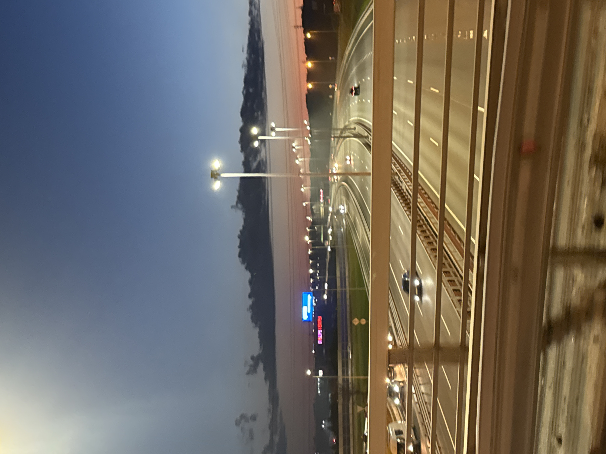 This is a vertical, eye-level, long shot photograph taken from a bridge overlooking a multi-lane highway at dusk. The sky is a gradient of deep blue at the top transitioning to a pale orange and pink near the horizon. Dark, wispy clouds are visible across the sky.
The highway below is illuminated by numerous streetlights and the headlights and taillights of vehicles. Cars and trucks are visible in various lanes, suggesting moderate traffic. The highway is bordered by concrete barriers and metal railings, part of which is in the foreground of the image, slightly out of focus. In the distance, there appears to be a large building with a blue illuminated sign and another sign with red lettering. The overall impression is a view of a busy urban highway at the end of the day, with the lights creating a dynamic and somewhat blurred scene. The location is identified as Korabsel'ki, Russia.