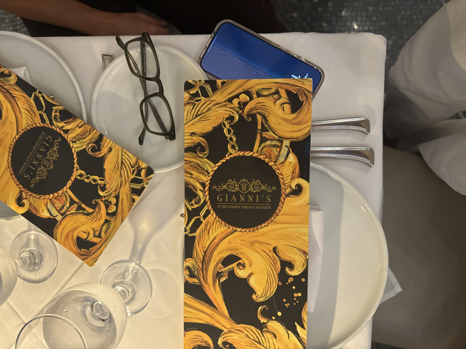 The image captures a dining table scene at Gianni's at The Former Versace Mansion, located in Miami Beach, United States. The table is draped with a white tablecloth. Two menus for "GIANNI'S AT THE FORMER VERSACE MANSION" are visible, featuring a black background with elaborate gold baroque patterns and chains. White ceramic plates are positioned on the tablecloth, accompanied by clear glassware, including a water glass and partially visible wine glasses. A pair of dark-framed reading glasses rests on one of the white plates. A smartphone with a blue screen/case is partially visible on the table. Silverware is arranged alongside the plates. A person's leg, clad in white trousers, is partially visible at the bottom of the frame, indicating an occupant at the table. A partial arm is also visible on the right side of the frame.