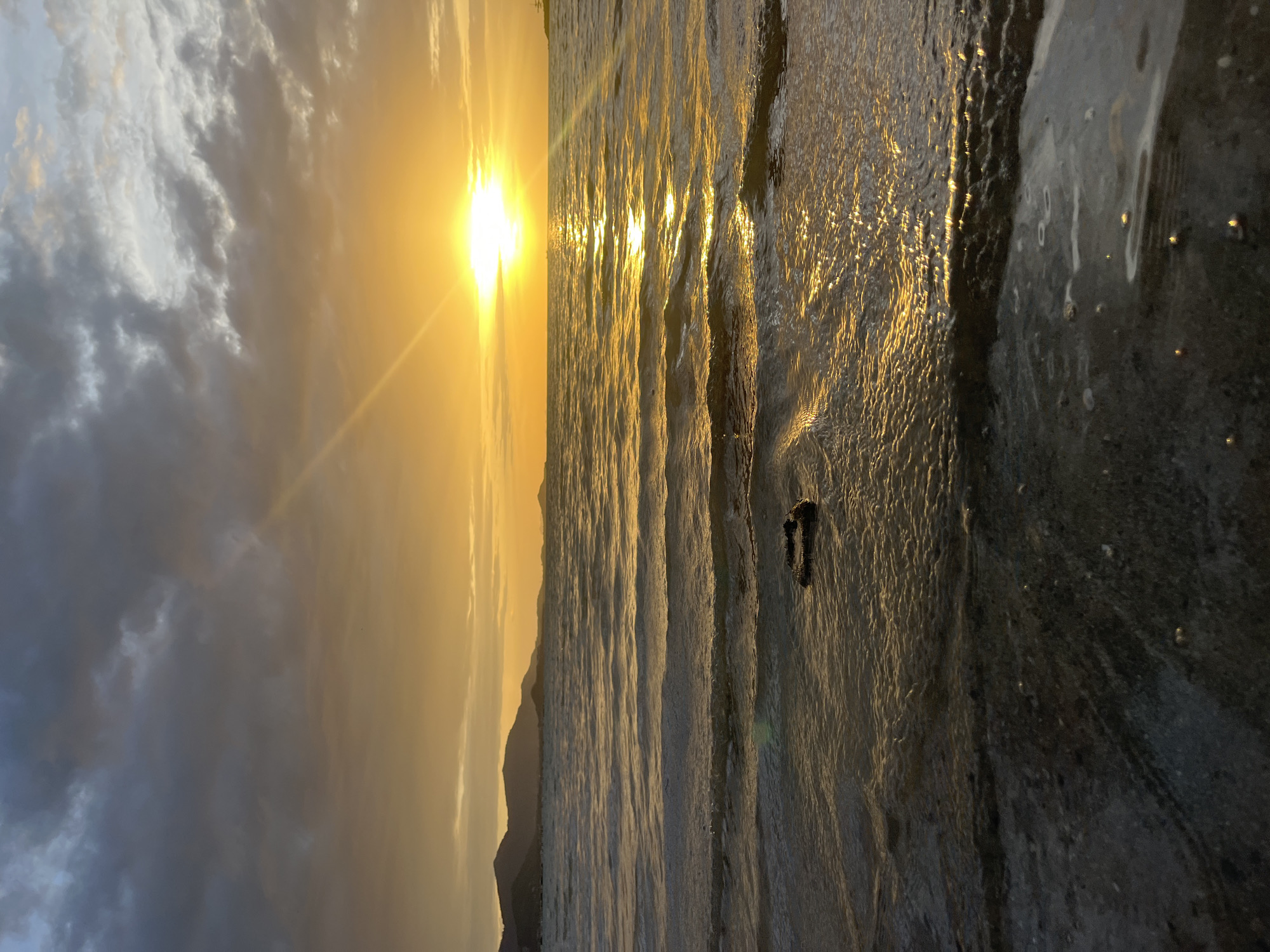 The image depicts a sunset over the ocean in Baan Plai Laem, Thailand. The sun, a bright orb of golden light, is low on the horizon, casting a brilliant glare across the rippling water. The sky is a gradient of warm oranges and yellows near the sun, transitioning to muted grays and blues as it ascends, with scattered clouds adding texture. In the distance, the silhouettes of rolling hills or mountains are visible against the vibrant sky. The foreground shows the shallow water lapping at a dark, pebbly shore. A small, dark object, possibly a piece of driftwood or debris, rests partially submerged in the water near the shore. The overall scene is serene and atmospheric, characteristic of a coastal sunset.