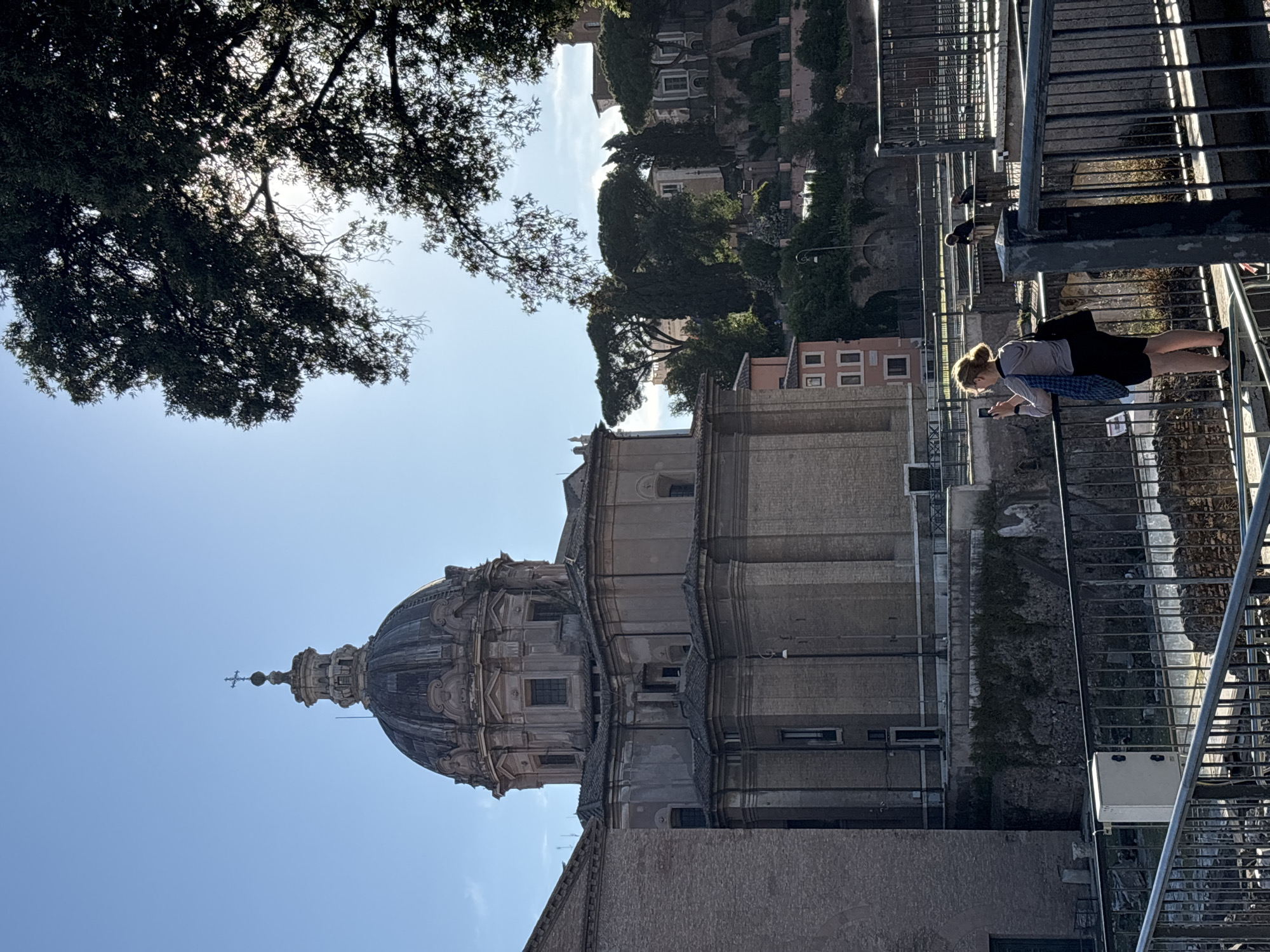 The image depicts a view in Rome, Italy, looking towards the dome of a large church. The dome is ornately decorated and has a cross atop its apex. A woman stands on a metal grated walkway, looking at a device. Two birds are nearby. The scene includes tree foliage and buildings.