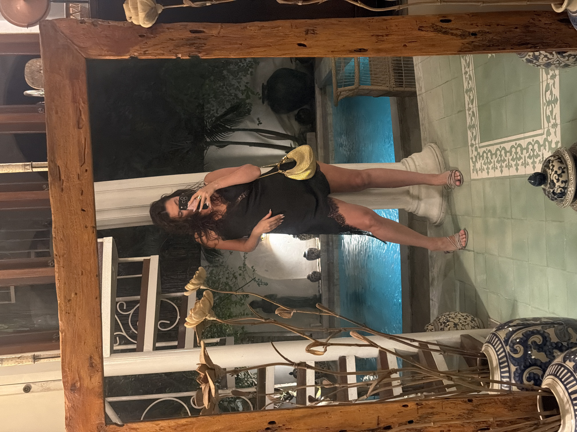 A woman stands in front of a large mirror, taking a selfie. She is wearing a black, asymmetrical dress with lace trim and a small gold purse. She is also wearing strappy sandals. The mirror reflects her and the surrounding environment, which includes a swimming pool, potted plants, and decorative vases. The scene appears to be an outdoor or semi-outdoor space, possibly a hotel or resort in North Kuta, Indonesia, given the tropical foliage and architectural style. The lighting suggests it is nighttime.