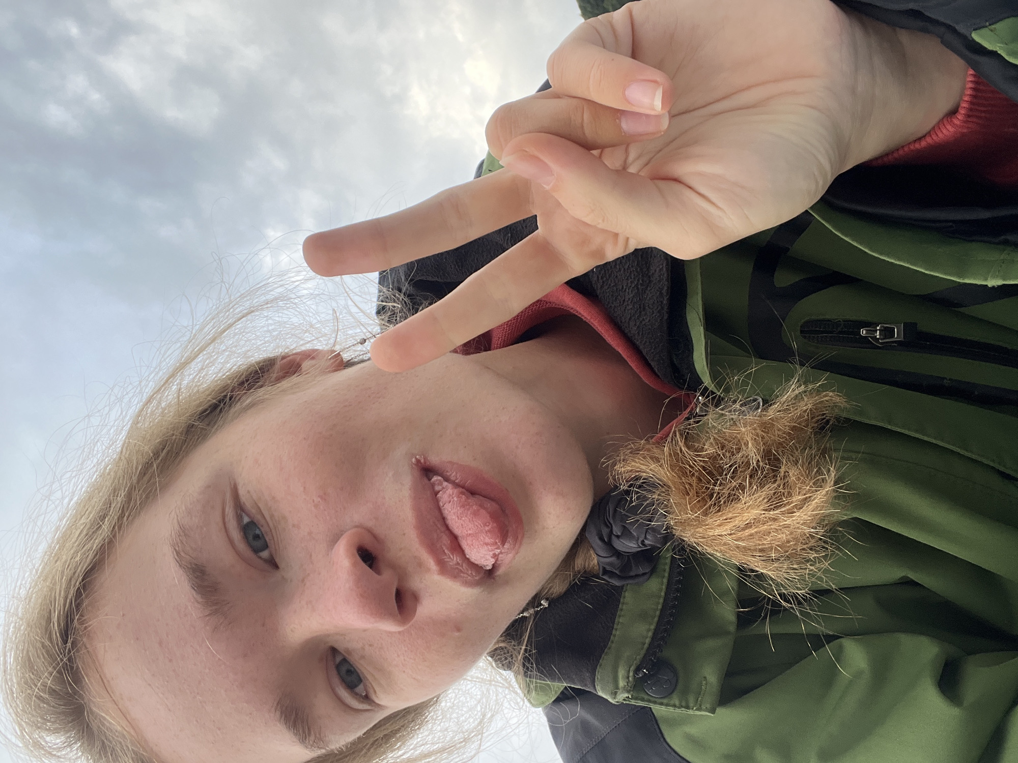 An angled close-up selfie of a person with blonde hair and fair skin, wearing a green and black jacket with a red underlayer, sticking their tongue out. The person is holding up a peace sign with their right hand. The background is a cloudy sky. It is not possible to confirm the location is in the city of Boreti, Montenegro from the image provided.