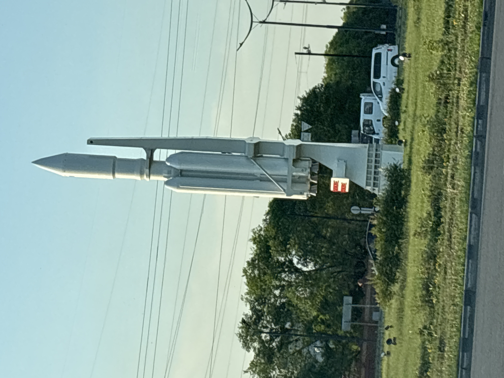 The image shows a large, white, multi-stage rocket replica displayed outdoors, likely as a monument or roadside attraction. The rocket is mounted on a white structure. The background includes a clear, light blue sky with power lines crisscrossing it. To the right of the rocket, there is a grassy area with trees and a road or sidewalk. Two white vans are parked in the grassy area, one behind the other. A few dark figures, possibly birds or small animals, are visible in the grass. The scene appears to be bathed in natural light, suggesting it was taken during the daytime, likely in the morning or late afternoon given the quality of the light. There are some signs visible, but no text is legible. The overall setting suggests an outdoor public space, possibly near a road or in a park area. The rocket's prominence suggests it is a significant local landmark.