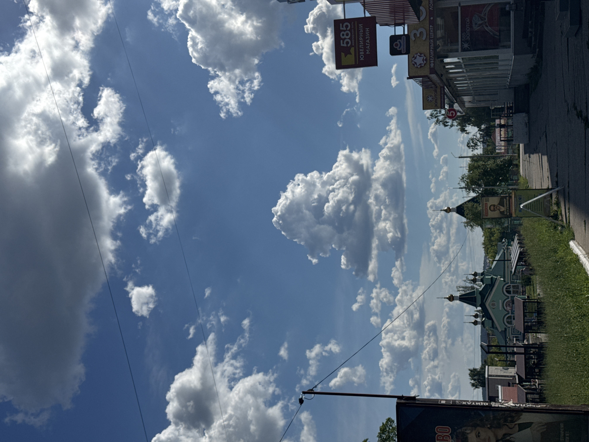 The image shows a street scene in Chaikovsky, Russia, on a bright, cloudy day. The camera is tilted, giving a slightly disorienting perspective. In the foreground, a dark billboard with an image of a woman's face and text partially visible reads "BO" and possibly Russian words related to "shield" and "limits". Further down the street, there are several shops with prominent signs. One sign clearly displays "585" with "ЮВЕЛИРНЫЙ МАГАЗИН" (Jewelry Store) and "ЮВЕЛИРНЫЙ ЦЕНТР" (Jewelry Center) in Russian, indicating a commercial area. Another sign says "3 * золотой" which likely refers to gold jewelry.
To the right of the shops, there is a sidewalk and a grassy verge. In the distance, a distinctive green building with golden domes, resembling a church or a cultural landmark, is visible. The sky is a vibrant blue, filled with large, fluffy white clouds, suggesting fair weather, possibly midday. Power lines crisscross the sky, adding to the urban feel of the scene. The overall impression is a typical Russian town street with shops, religious architecture, and a bright, cloud-filled sky.