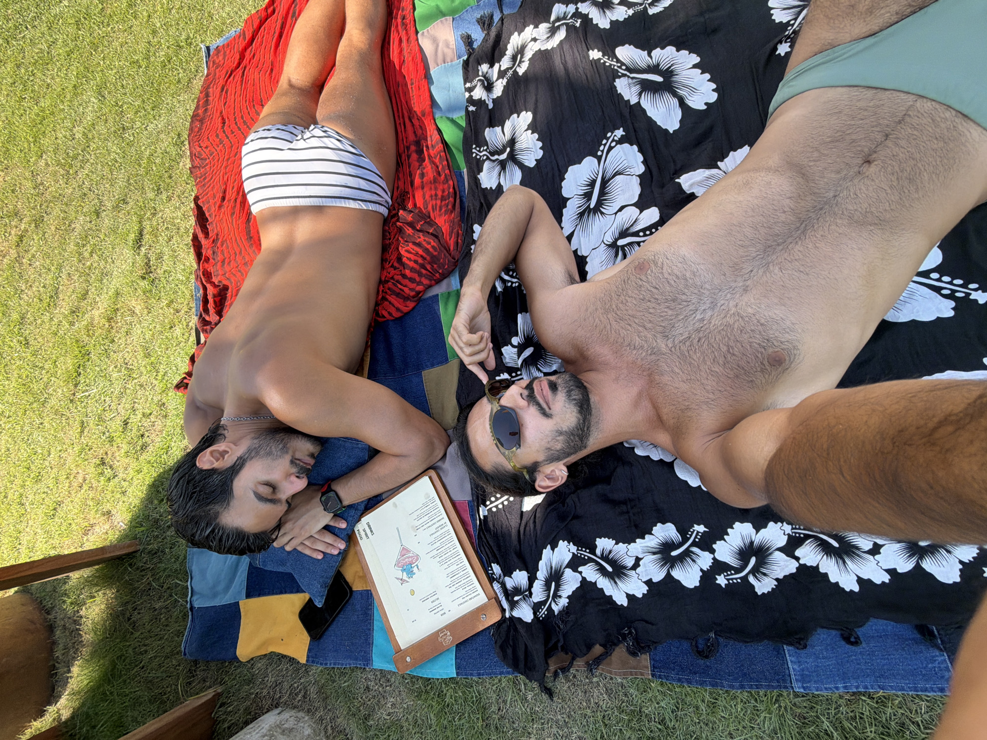 Two men are lying on a patchwork quilt blanket on grass outdoors, possibly in Mengwi, Indonesia, given the provided location. One man is lying on a red and black patterned cloth, wearing striped swim trunks, a watch, and a necklace, with his eyes closed. The other man, wearing sunglasses and green swim trunks, is lying on a black cloth with white hibiscus flowers, gesturing with his finger. A restaurant menu and a phone are resting on the blanket near the men.