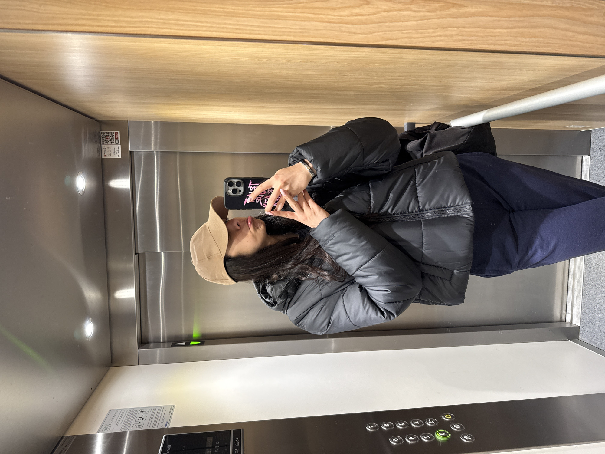 A person in a black puffer jacket and a tan baseball cap is taking a selfie in an elevator. The elevator has a brushed metal interior with a wood panel above. A white handrail runs horizontally along one wall. The elevator's control panel with floor buttons is visible at the bottom of the image. The location is Sofia, Bulgaria.