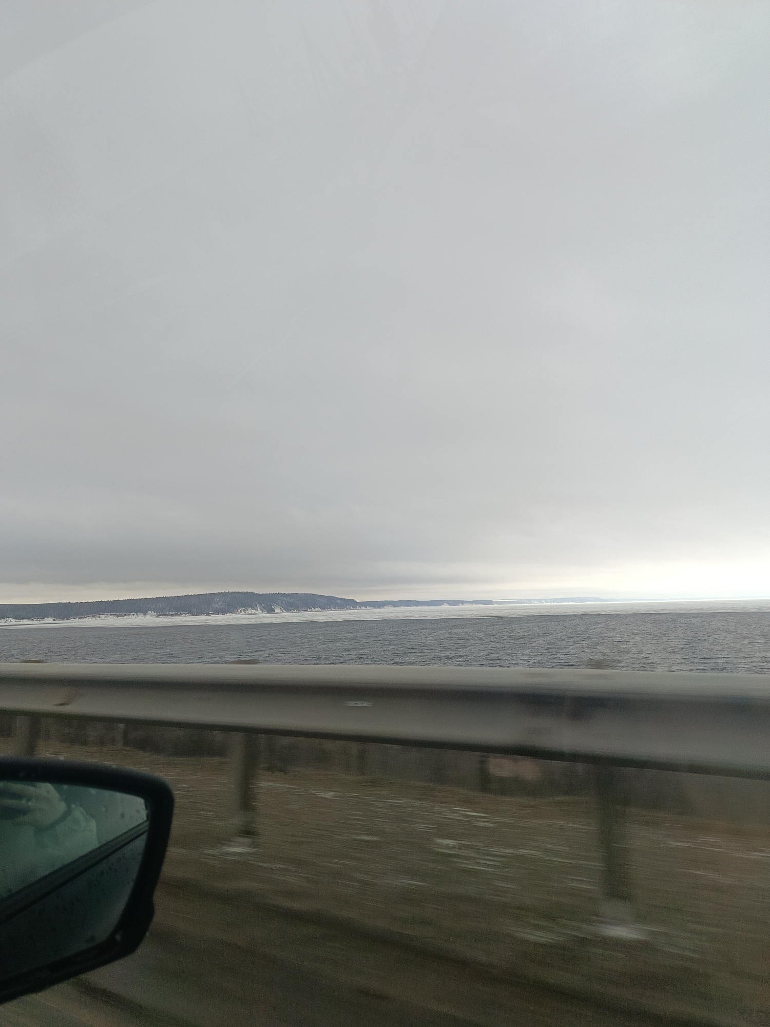 The image shows a landscape view taken from a moving vehicle on a bridge or elevated road, likely in or near Chaikovsky, Russia, based on the landscape and infrastructure.
The foreground includes the vehicle's side mirror and a portion of the guardrail. The road surface appears blurred due to motion.
The midground features a body of water, possibly the Kama River, with a slightly rippled surface and some ice or snow cover near the shoreline. In the distance, there's a landmass, possibly an island or the opposite bank of the river, covered with trees, some of which appear to be snow-covered.
The background is dominated by a gray, overcast sky. The overall lighting is muted, suggesting a cloudy or winter day. No people are visible in the frame.