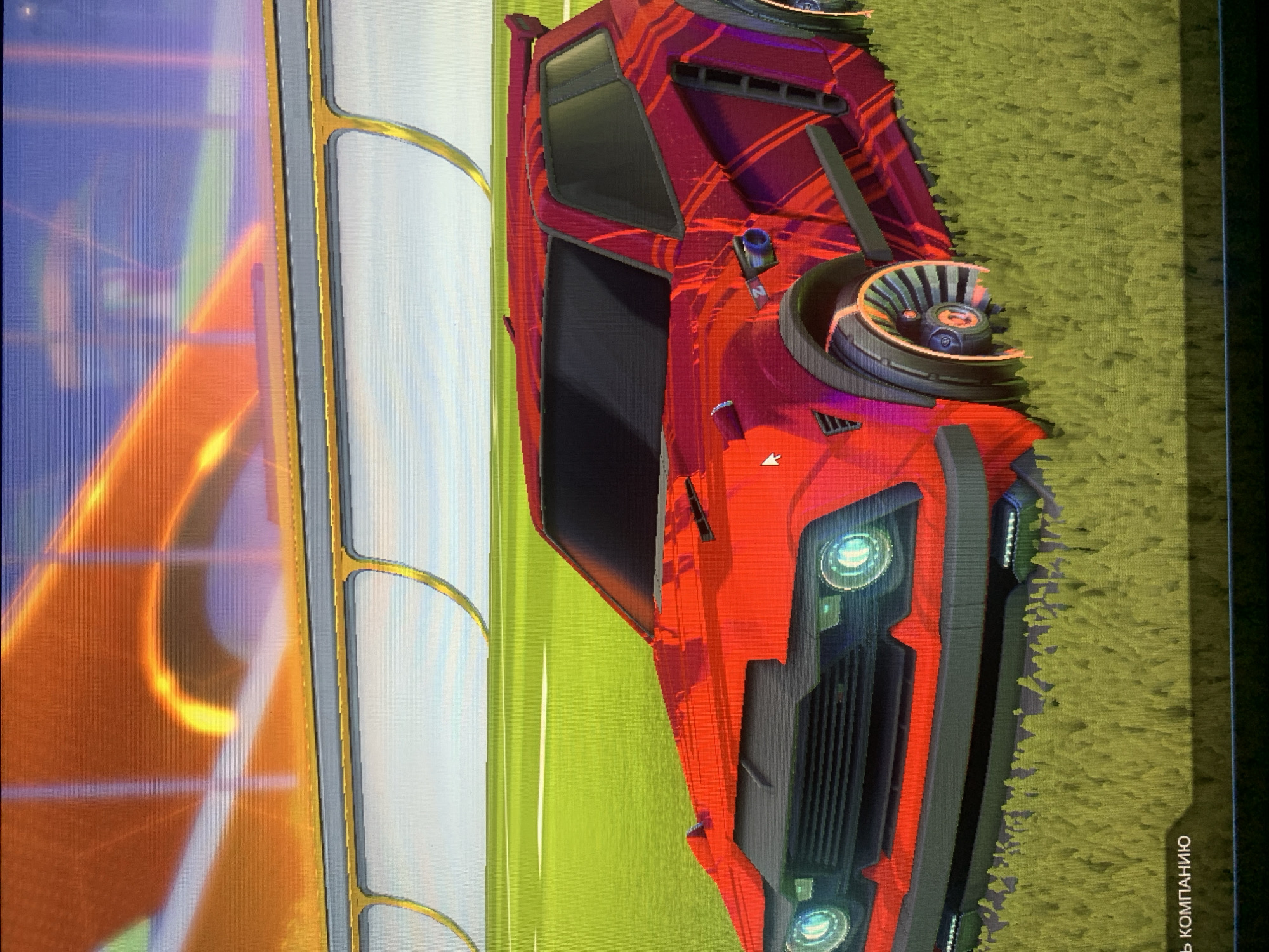 A red car with a red and orange striped decal is parked on a bright green field. The car has blacked-out windows and silver wheels with orange accents. In the background, there is a stadium with a yellow railing and an orange and yellow illuminated structure. The bottom of the image shows a portion of text in Russian that translates to "company". The scene is likely from a video game, possibly Rocket League, given the car design and stadium environment.