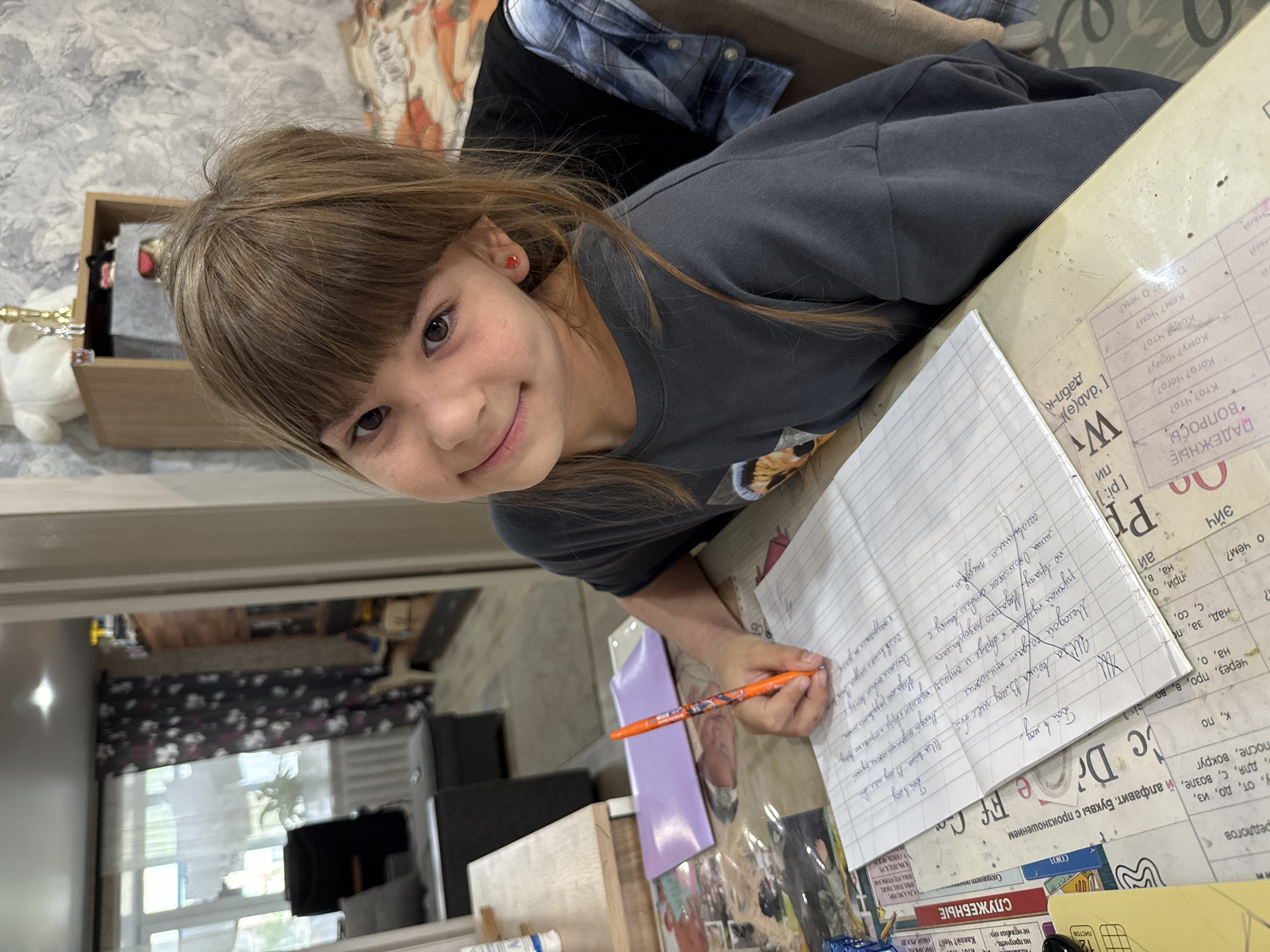 A young girl is sitting at a table, writing on a sheet of lined paper with an orange pen. She is wearing a gray t-shirt and has brown hair with bangs. She is smiling and looking at the camera. The table is covered with papers and other objects, including a purple notebook. In the background, there is a window with sheer curtains, a television, and a dresser. It is difficult to determine the exact location in Rusanovo, Russia without additional context.
