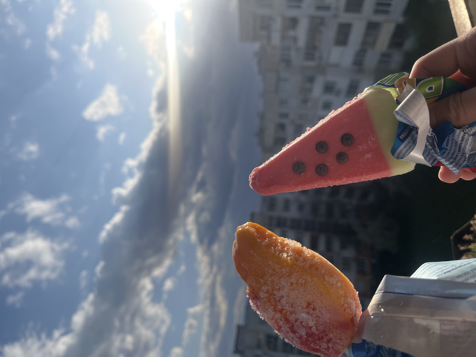 The image shows two ice cream pops being held up against a bright sky with clouds and a building in the background. One ice cream pop is watermelon-shaped, with red, white, and black colors. It is being held in a person's hand with part of the wrapper visible. The other ice cream pop is orange and red. It has a frosted appearance, suggesting it is very cold. The background includes a building and a bright sky with scattered clouds and a bit of sun glare. Based on the architecture, the photograph might have been taken in Sofia, Bulgaria.