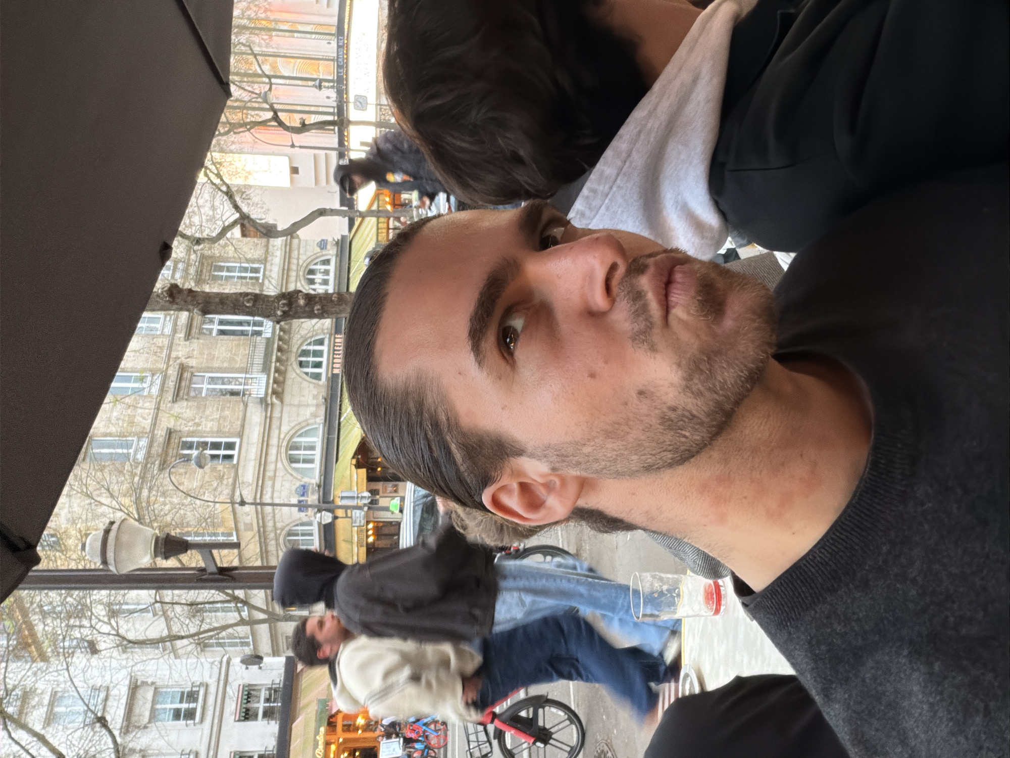 A close-up portrait shows a man with dark hair and a beard. He is wearing a dark gray sweater. His gaze is directed towards the camera. Behind him, the blurred background shows a street scene with pedestrians and buildings typical of Paris, France. A couple is on a bicycle ride. Details include buildings with multiple windows and architectural elements, lampposts, and trees with bare branches, all consistent with a Parisian cityscape. The picture is taken from under a dark umbrella at an outdoor terrace.