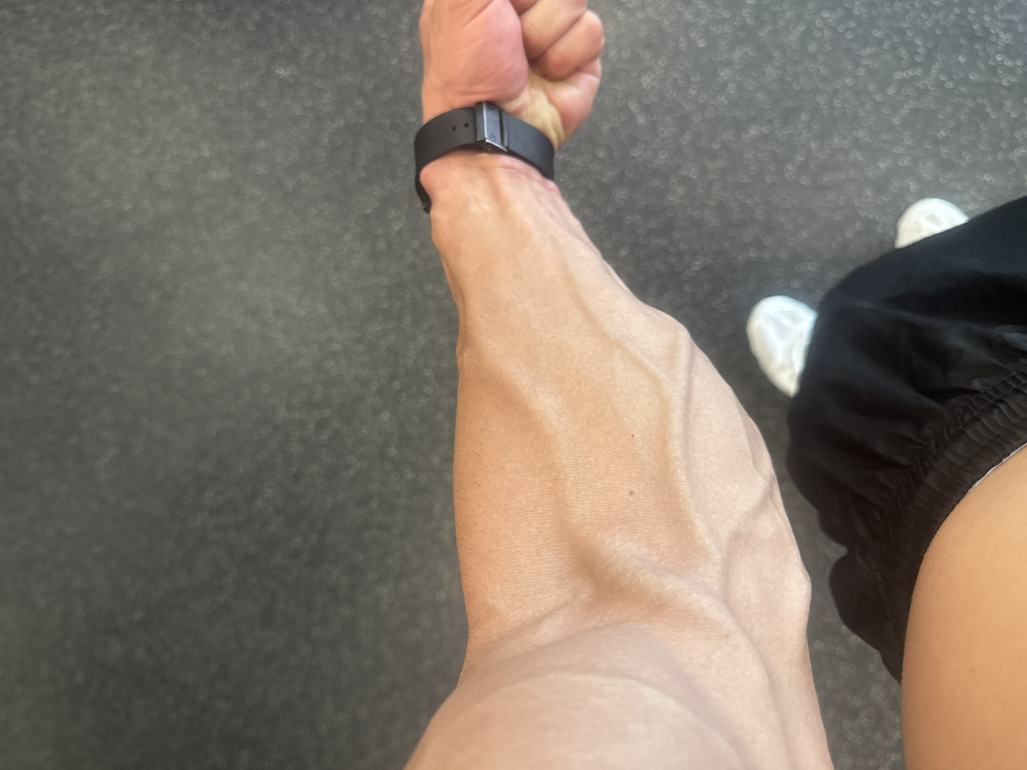 The image shows a person's arm and wrist with a black smartwatch. The arm appears muscular with prominent veins. The person is wearing black shorts and white shoes. The background is a gray, textured floor.