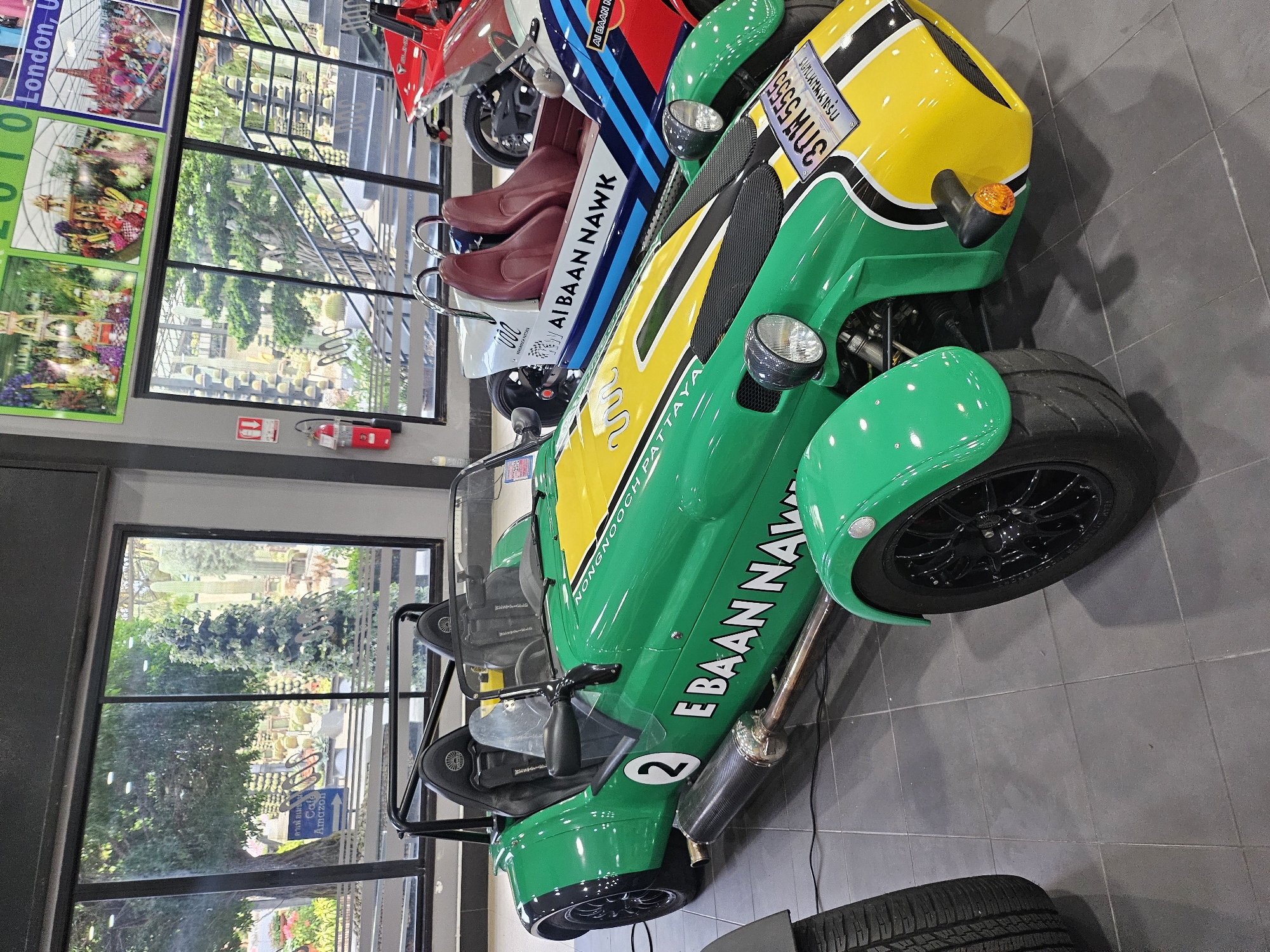 The image shows a bright green, open-top sports car with a yellow stripe, parked indoors on a gray tiled floor. The car has a roll cage, two seats, and the number "2" on its side. Text on the car reads "E BAAN NAWK" and "NONG NOOCH PATTAYA". A license plate on the front reads "3กพ 5555". Behind the green car, another sports car with red seats and "AI BAAN NAWK" text is visible, followed by a red motorcycle. Large windows in the background reveal a garden setting with many cacti and lush greenery, and a sign pointing to "Cafe Amazon". The overall environment appears to be a showroom or a visitor center.