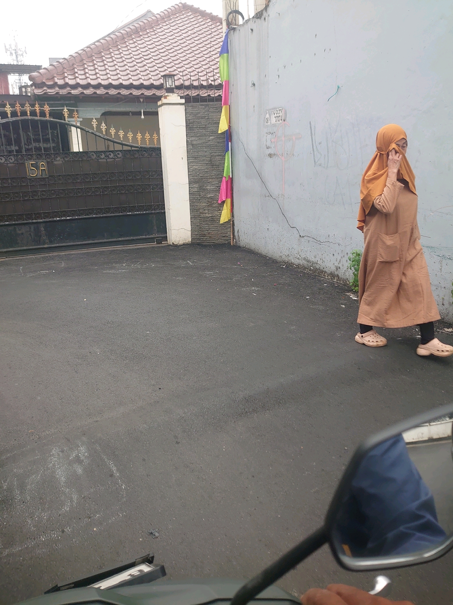 A woman wearing a light brown hijab and a matching long, loose dress walks along a paved street next to a textured gray wall. The woman is positioned on the right side of the frame, partially obscuring her face with her hand. A black metal gate with ornate gold accents, labeled "15A", is visible on the left side of the image, in front of a house with a reddish-brown tiled roof. Colorful small flags hang from the wall behind the gate. The street appears wet, suggesting recent rain. The foreground of the image shows a portion of a motorcycle, including a side mirror reflecting a blue garment. This scene is located in Depok, Indonesia, a city known for its residential areas and urban development.