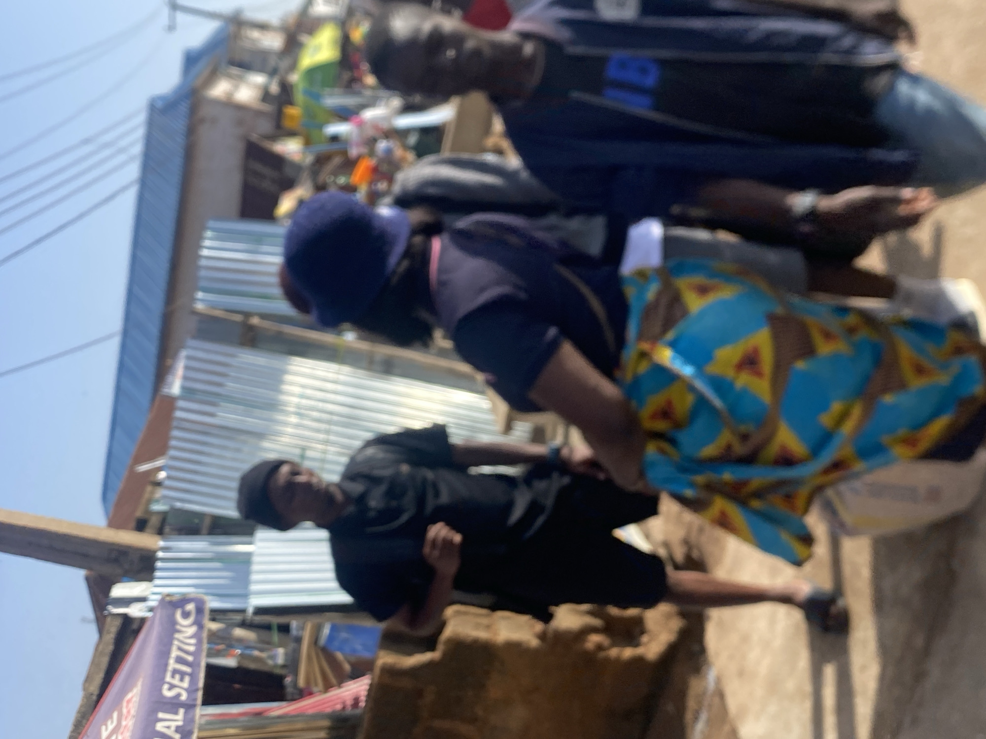 In Bukuru, Nigeria, a street scene unfolds under a bright sky. The foreground is dominated by four individuals. On the left, a person in a black polo shirt and black shorts stands with their left hand clenched in a fist. Their face is partially obscured by a black beanie. To their right, a person wearing a dark blue hat and a dark blue t-shirt with a brightly colored blue, yellow, and red patterned fabric wrapped around their waist is partially visible, facing away from the camera. Behind them, another person, wearing a dark blue jacket over a black t-shirt with blue lettering that appears to spell "IB", is seen from the chest up, looking towards the right. In the far right background, a fourth individual is partially visible. The environment includes corrugated metal sheeting and a building with a blue roof. A purple awning with white lettering that reads "AL SETTING" is visible on the left side of the image. The ground appears to be dirt or sand. The overall impression is a busy outdoor market or street environment.