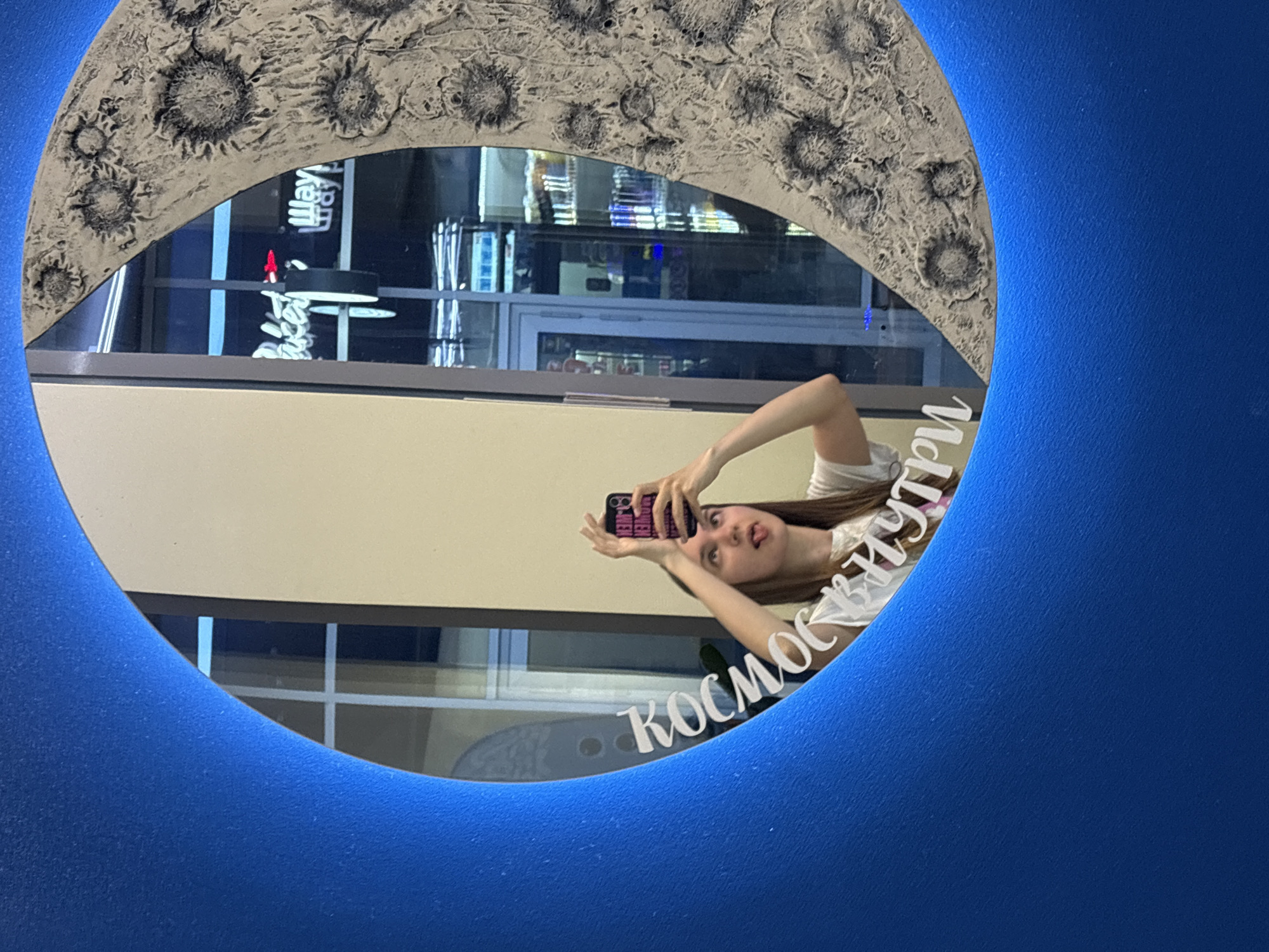 A young woman with long hair is reflected in a circular mirror with a lunar surface pattern around the edge and blue framing. She is holding a smartphone, presumably taking a selfie. The reflection also reveals an exterior scene, possibly a building with illuminated signs and window panes. The text "КОСМОС ВНУТРИ" (translated: "Space Inside") is written in white on the mirror surface. Based on the mirror's design and available context, it's plausible this photo was taken in the city of Perm, Russia.