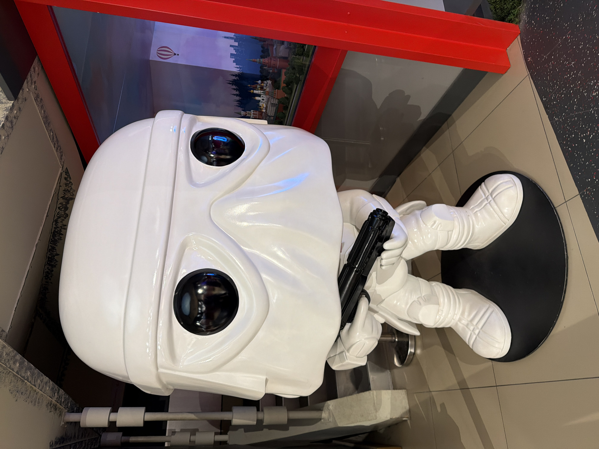 A large, glossy white figurine resembling a Star Wars Stormtrooper is positioned indoors. The figure, a stylized representation with large, black, round eyes and holding a black blaster, stands on a black circular base on a tan tiled floor. A red structure frames a backdrop showcasing a cityscape of Moscow, Russia, complete with recognizable landmarks and a hot air balloon in the sky. The figurine is placed near a wall and adjacent to what appears to be another structure or display.