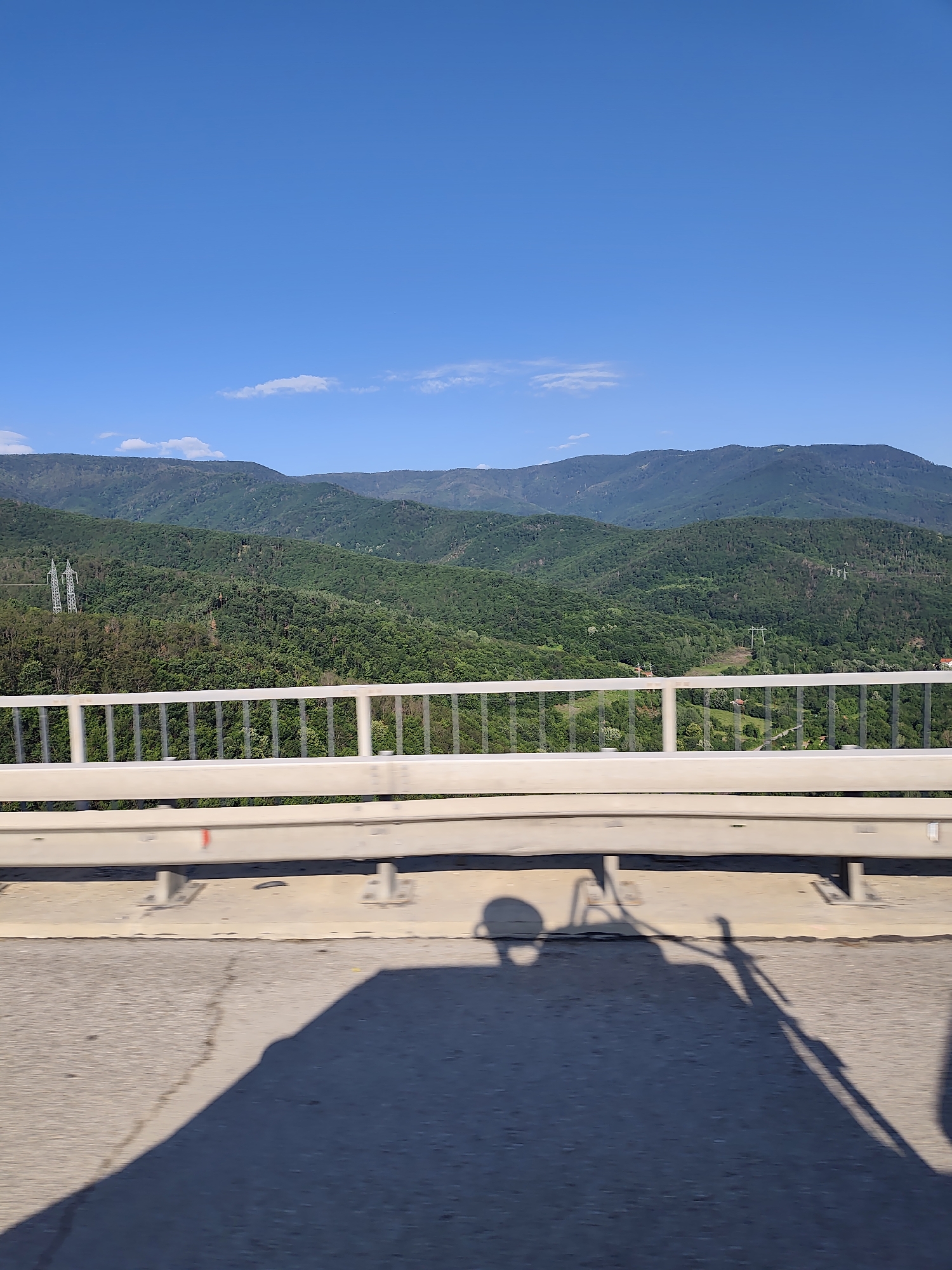 A sunny day over a lush, green mountain range in Vrachesh, Bulgaria is depicted from a bridge. The foreground features a concrete road surface with distinct shadows of people cast on it, suggesting movement. A white metal guardrail runs horizontally across the mid-ground, separating the road from the expansive vista. Beyond the guardrail, rolling hills covered in dense green forest stretch into the distance, with a clear blue sky dotted with a few wispy clouds overhead. A single, tall electricity pylon is visible on one of the forested slopes. The overall impression is one of natural beauty and a bright, clear day in a mountainous region.