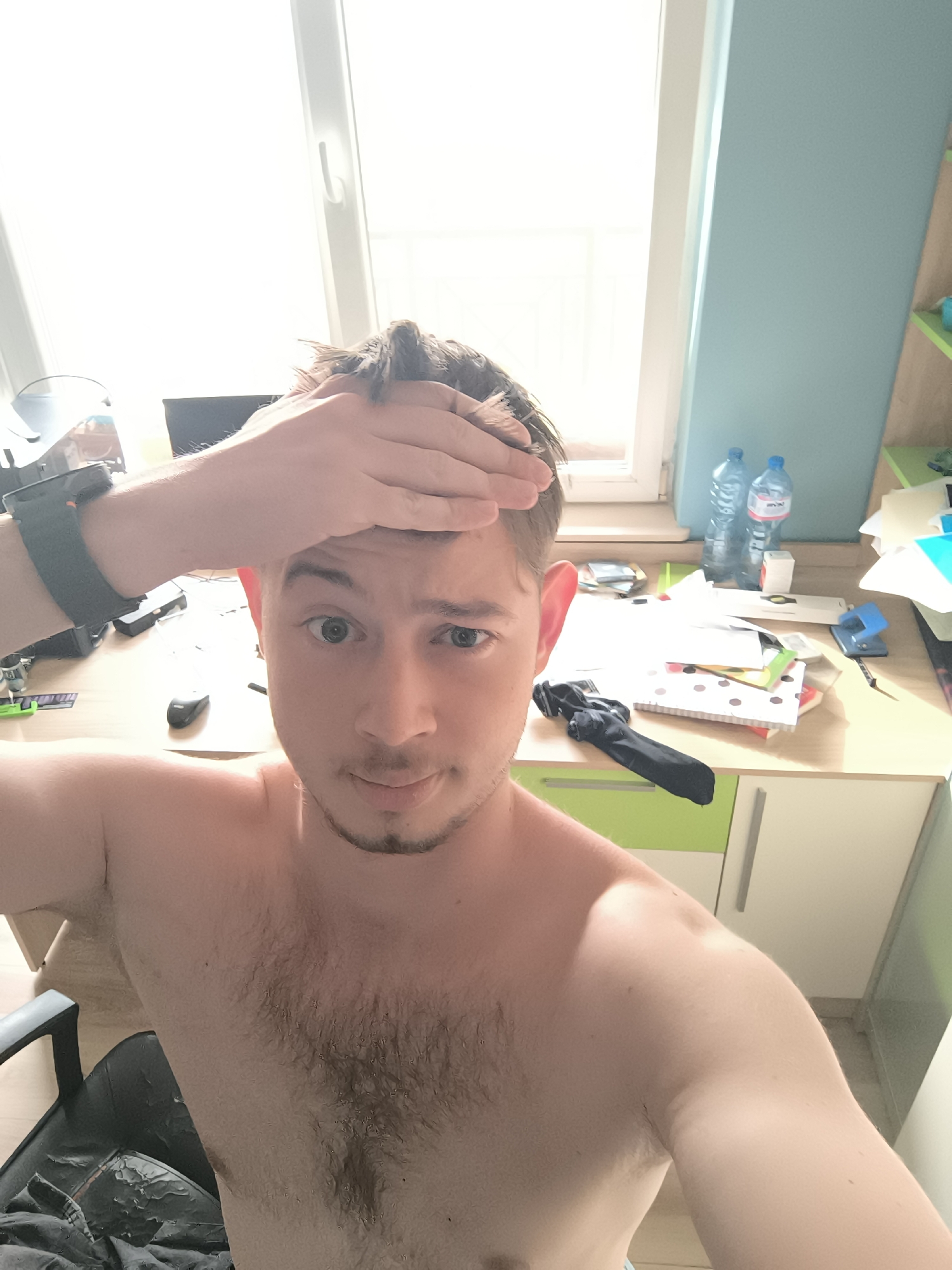 A shirtless fair-skinned male is taking a selfie indoors. He is holding his right hand to his forehead. He has short brown hair and light eyes. He has a watch on his left wrist. Behind him is a cluttered desk with two water bottles, papers, and office supplies. There is a window with bright light coming through and a blue wall in the background. A chair is visible on the left side of the image. The location is not discernible as being in Sofia, Bulgaria.