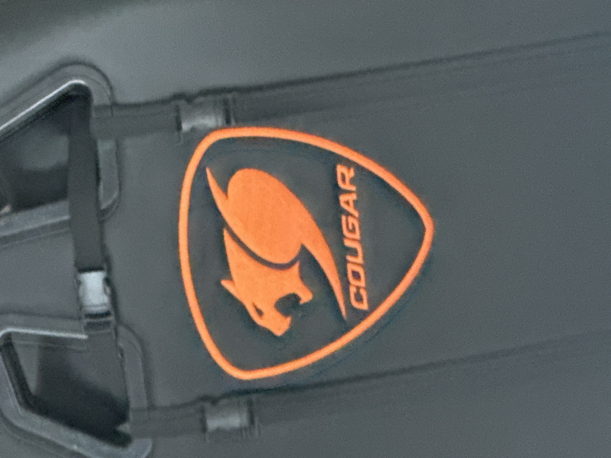 The image presents a close-up, static view of a dark grey, slightly textured surface, likely fabric or molded plastic. Centrally positioned is an embossed or embroidered orange logo for the brand "COUGAR." The logo features a stylized cougar or mountain lion in an aggressive, pouncing stance with its mouth open, enclosed within an orange shield-shaped outline. Below the animal graphic, the brand name "COUGAR" is rendered in orange, capitalized block letters. Above the logo, dark grey webbing straps are visible. One strap runs horizontally across the top, featuring a small, light grey plastic side-release buckle. Another dark grey strap appears vertically, connecting to a larger, dark grey, curved or U-shaped structural element at the top of the frame. No individuals, specific actions, or environmental cues are visible to ascertain the precise nature of the object or its surrounding location. The image does not contain any information to indicate a location in Saraia, Bulgaria, or any other specific geographical area.