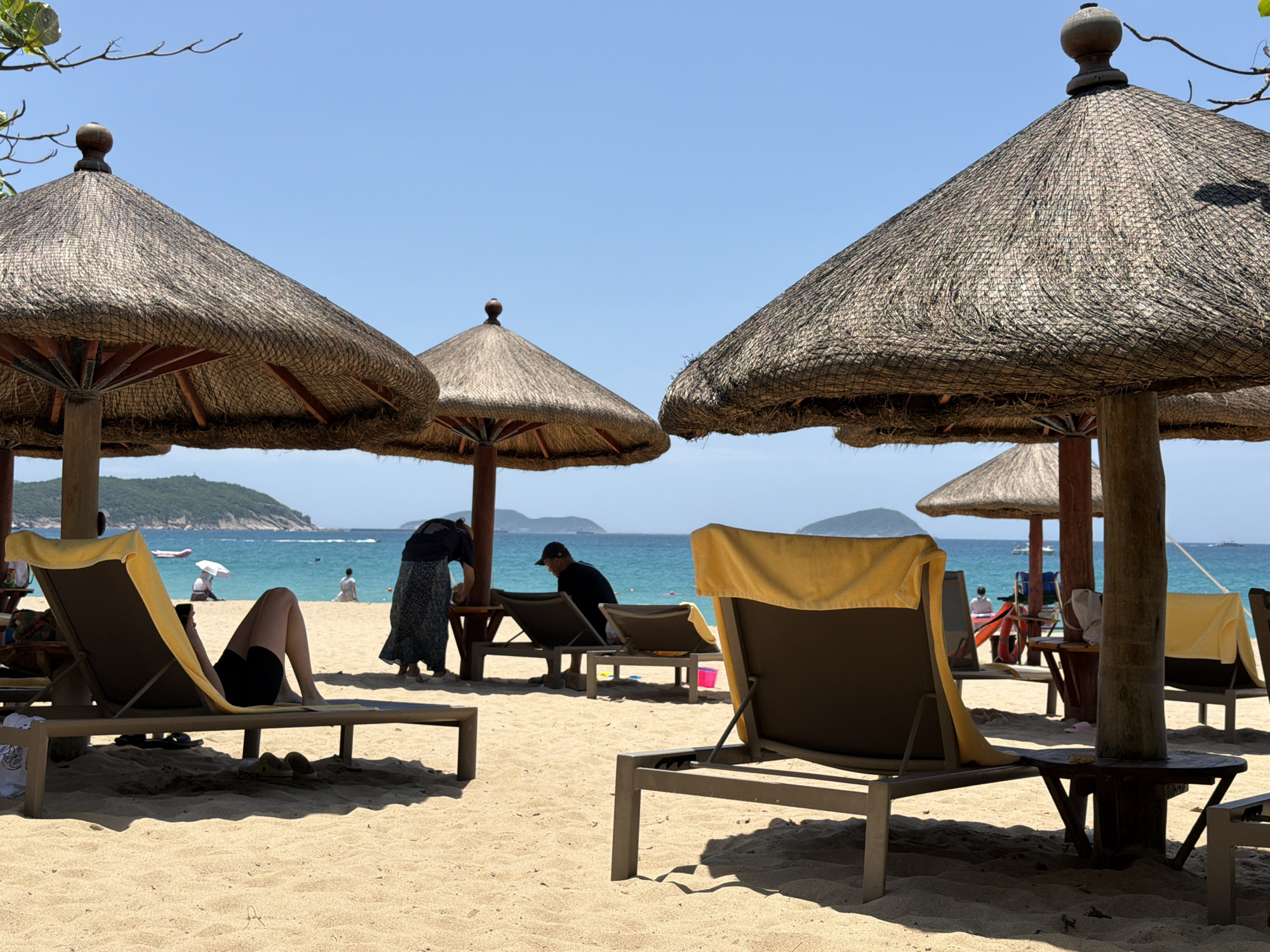 In Jiyang District, China, a serene beach scene unfolds under a clear blue sky. The foreground is dominated by a sandy beach with several thatched umbrellas casting shadows. Lounging chairs, some with yellow towels draped over them, are arranged on the sand. In the middle ground, two people are visible. One, wearing a black dress, is bending down near a lounge chair. Another person, wearing a black baseball cap and shirt, is seated on a lounge chair, facing away from the camera. Further out, the turquoise ocean stretches towards a distant, lush green island. A few other beachgoers and small boats are visible in the water. The overall atmosphere is one of relaxation and leisure in a tropical setting.