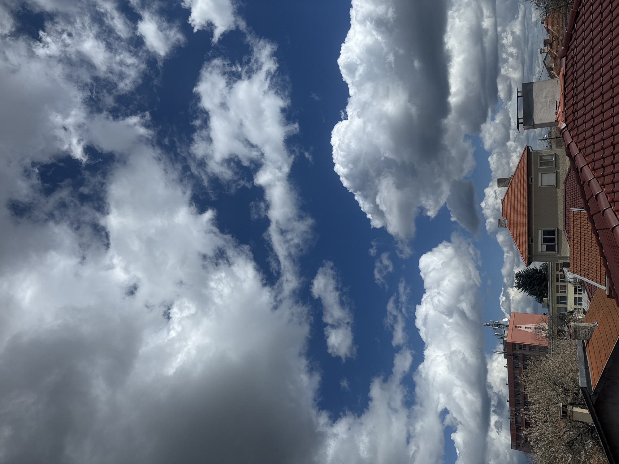 The image shows a daytime view of the sky and rooftops in Dobrich, Bulgaria. The sky is mostly blue with white cumulus clouds, with some areas obscured by darker, gray clouds. In the foreground, there are several buildings with red tile roofs, and some with gray facades. An apartment building with antennae on the roof is visible as well, indicating an urban location. Trees with sparse foliage are also present.