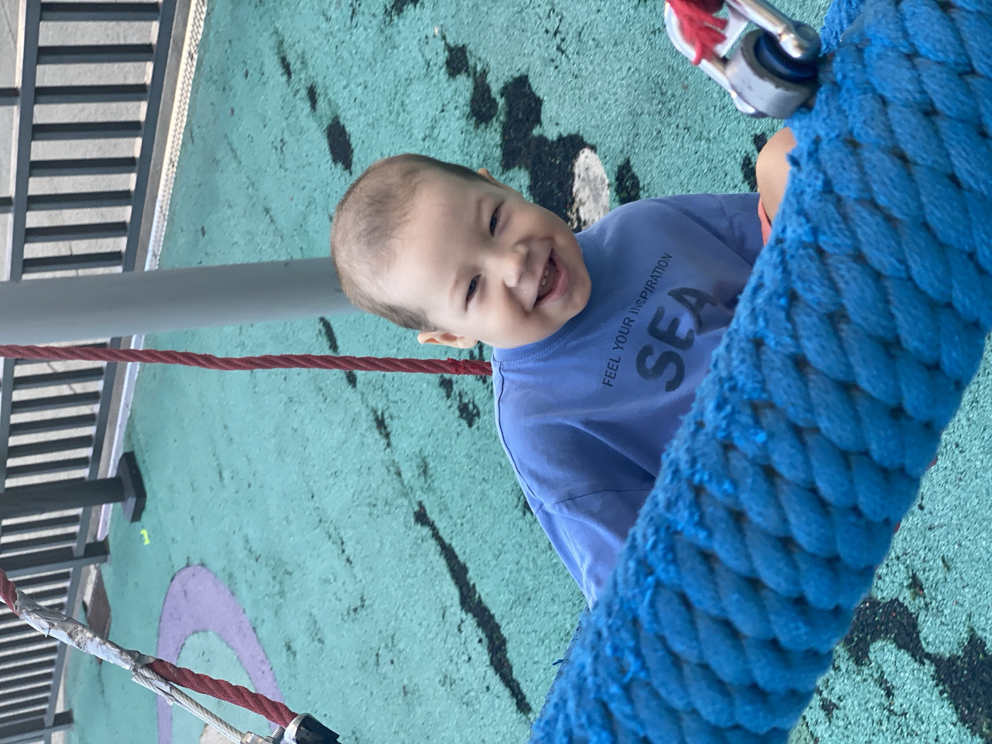 A young boy with light brown hair and a wide smile is sitting on a playground, possibly on a swing or zipline, as a thick blue rope is in the foreground. He is wearing a light blue t-shirt with the words "FEEL YOUR INSPIRATION SEA" printed on it. The playground surface is a textured teal color with purple markings and black streaks. In the background, a gray metal fence is visible, and the scene appears to be outdoors. The location is identified as Sochi, Russia.