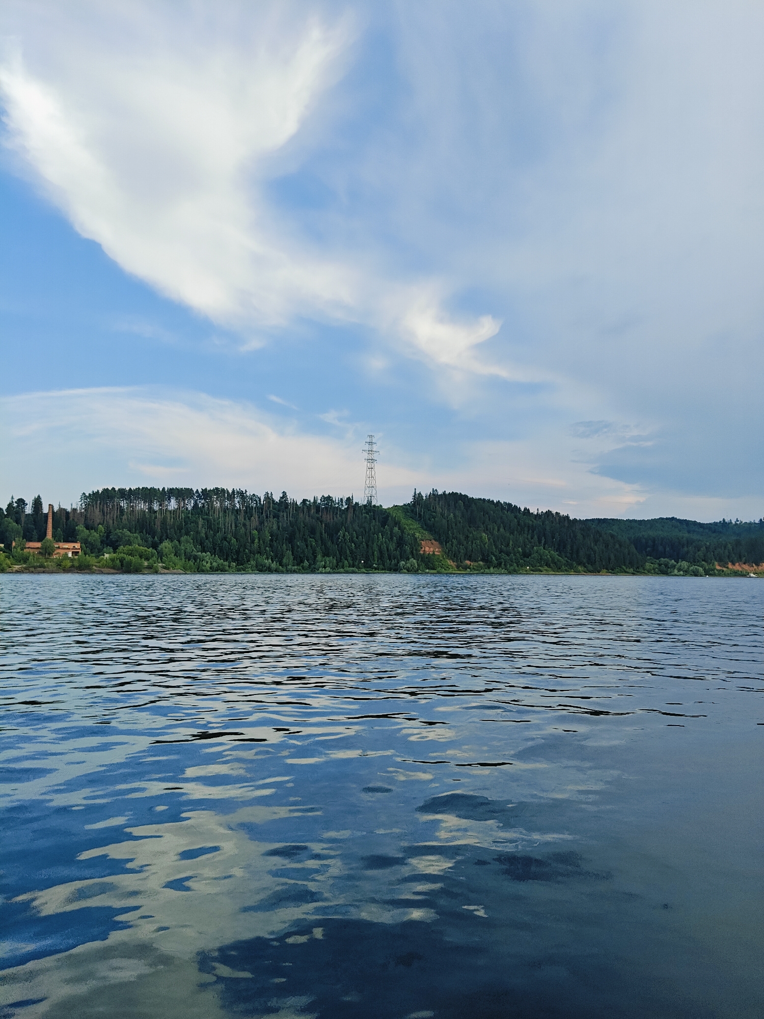 The image depicts a wide body of water, likely a river or lake, with ripples disturbing its surface, reflecting the sky and surrounding landscape. On the far bank, a dense forest covers rolling hills. A prominent electrical pylon stands tall on the right side of the forested hill, and to the left, a brick structure, possibly an old industrial building or tower, is visible amongst the trees. The sky above is a mixture of blue and white clouds, with a large, wispy cloud formation dominating the upper left quadrant. The scene appears to be a natural landscape with some human-made infrastructure. The location is identified as Noviy, Russia.