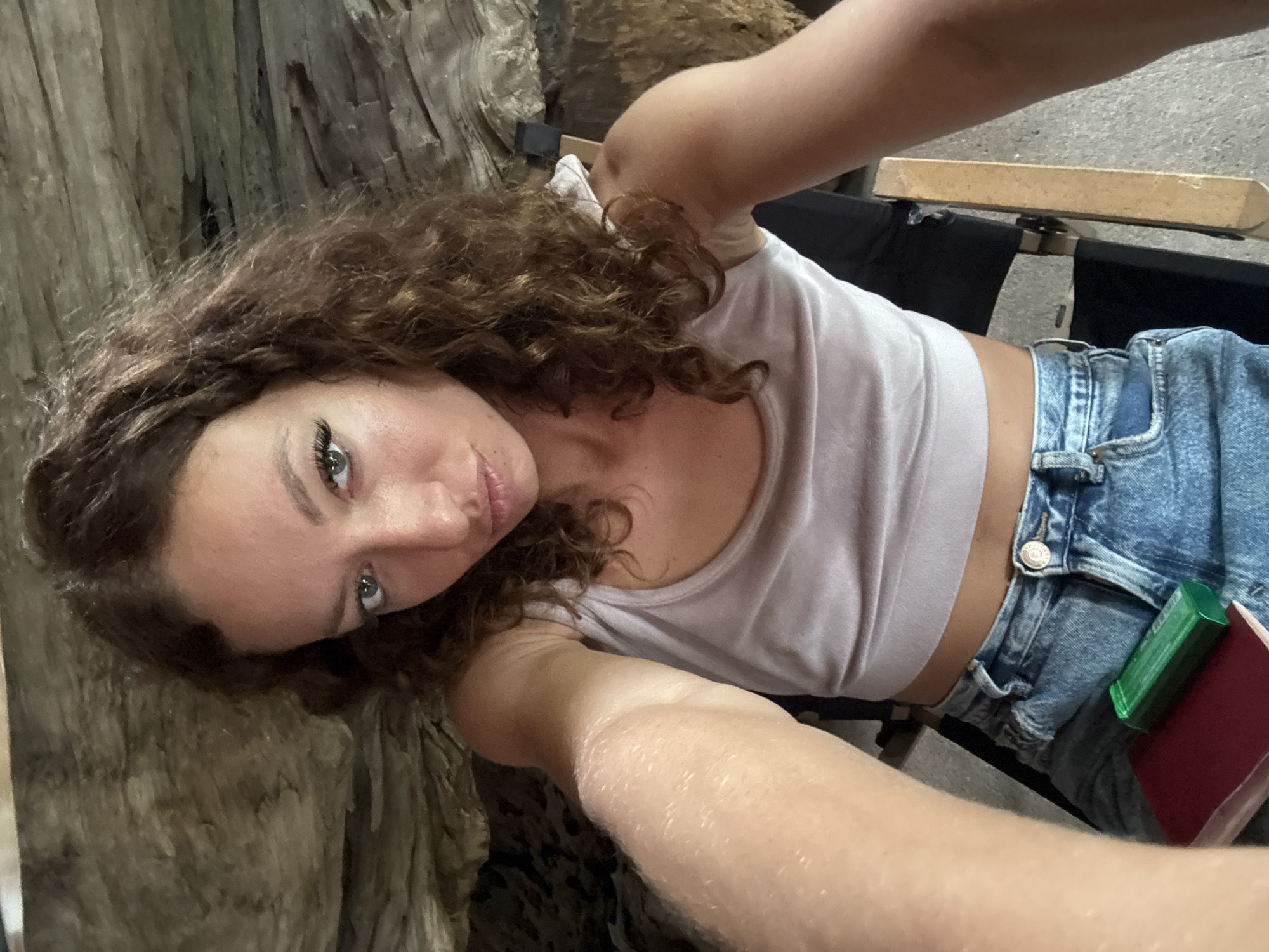 A selfie taken by a young woman with curly brown hair and light eyes. She is wearing a light-colored tank top and blue jeans. A green and red item is visible tucked into the waistband of her jeans. She appears to be sitting on a black director's chair with a wooden armrest. Behind her is a textured, gray, weathered surface, possibly a tree trunk or wooden structure.
While I can describe the image in detail, I have no information to suggest the location is in the city of Bản Đêu, Vietnam.