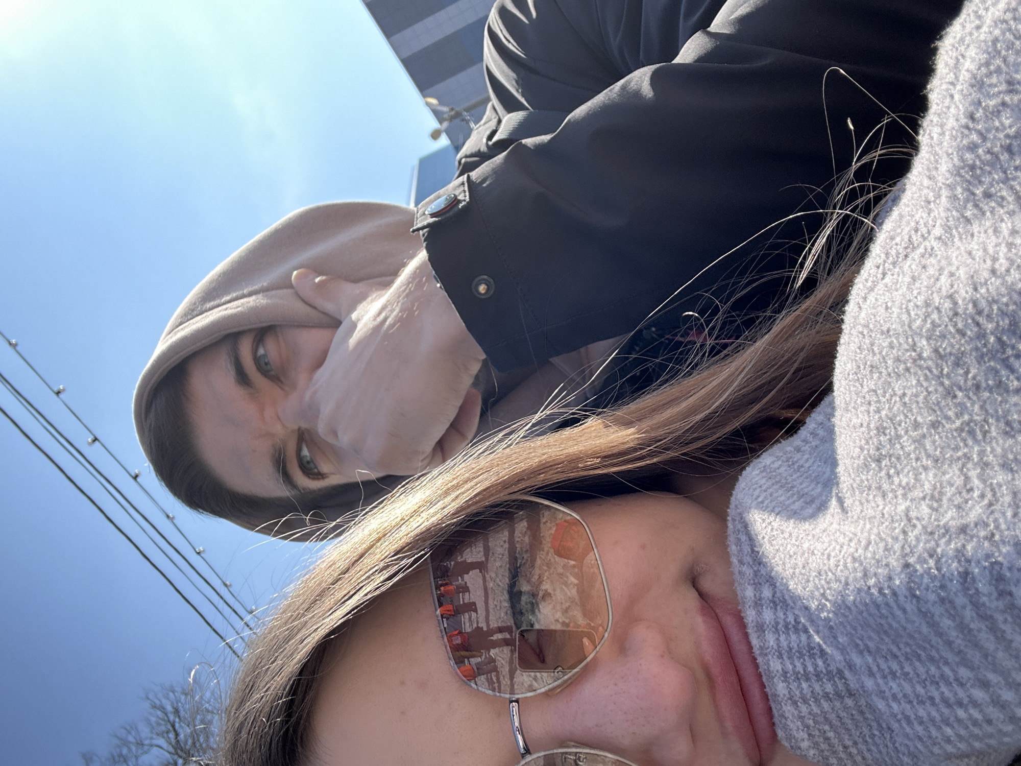 A selfie photograph, angled upward, depicts two individuals outdoors. The lower person, primarily visible from the nose and mouth up, is wearing sunglasses that reflect a scene of an outdoor event in the lenses, with red figures and structures. A portion of a gray knitted garment is also in view. Above them, the other person, wearing a hooded beige jacket with a hand partially covering their mouth, stares directly into the camera with a scowling expression. A black jacket is also visible. The background reveals a blue sky, power lines, bare tree branches, and the corner of a tall building. The location is in a city with architectural style similar to Zelenodol'sk, Russia.
