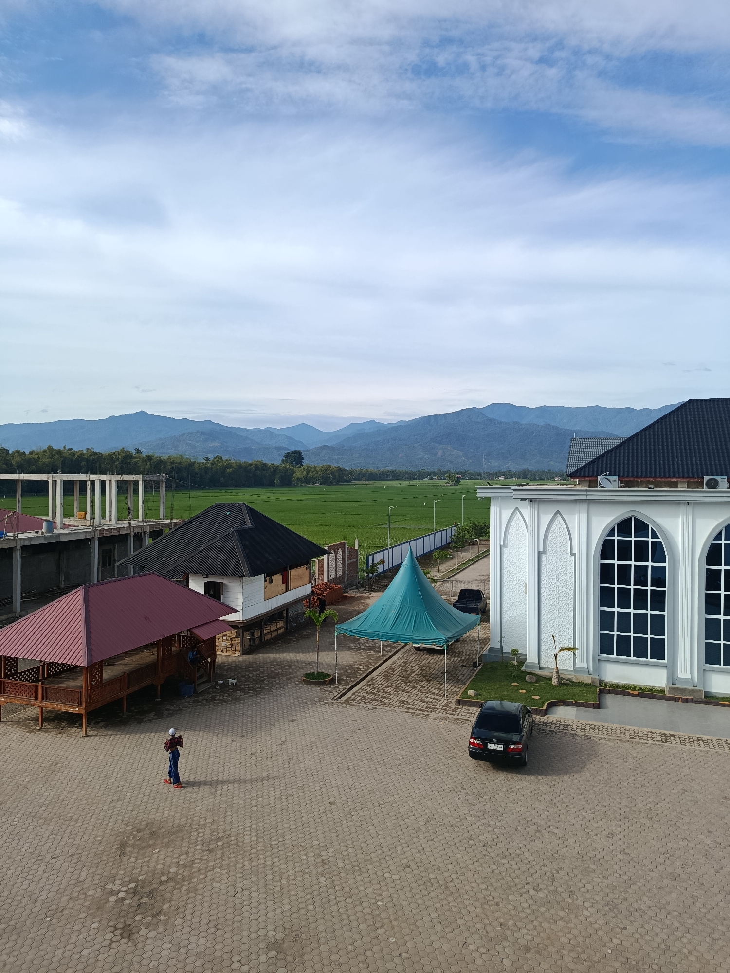 The image depicts an outdoor scene with a backdrop of lush green rice fields and distant mountains, under a partly cloudy sky. In the foreground, a paved area with hexagonal stones serves as a courtyard. Several structures are visible: a white building with arched windows and intricate decorative panels, a dark-roofed structure with white walls and a raised foundation, and a red-roofed open-air pavilion with wooden supports. A teal-colored tent is pitched in the courtyard. A black sedan is parked on the pavement. A person wearing a blue shirt, dark pants, and red shoes stands in the courtyard, facing away from the viewer. Construction is underway on a multi-story building with concrete pillars on the left side of the image. The location is identified as Peulakancibrek, Indonesia.