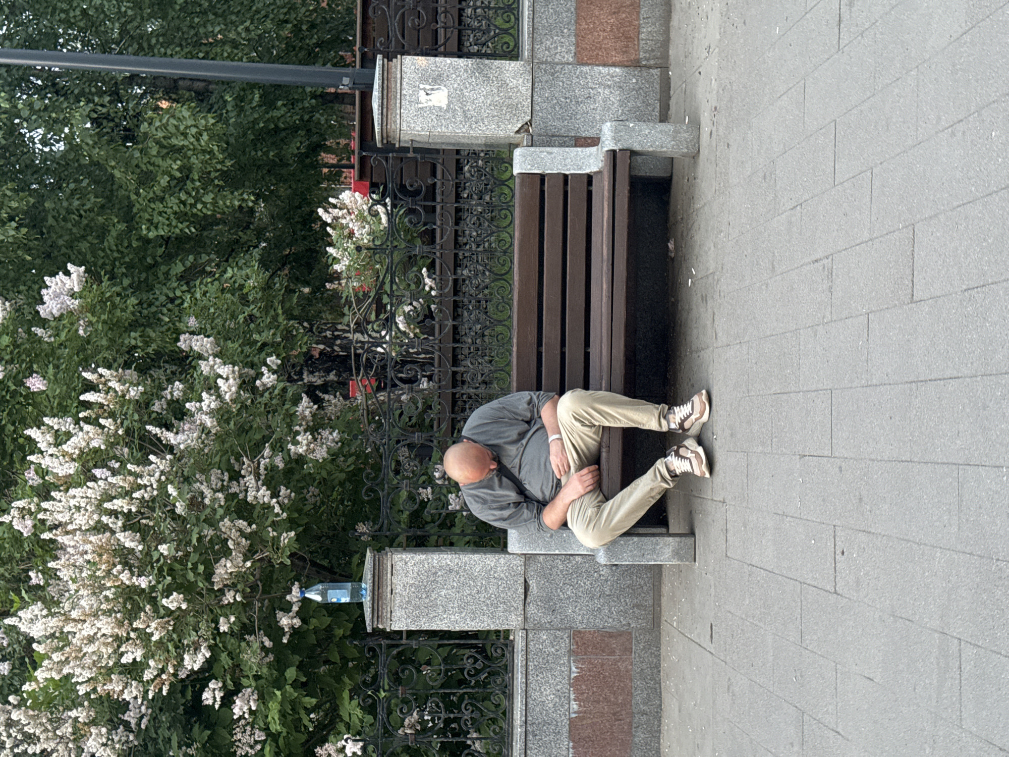 A bald, middle-aged man wearing a grey t-shirt and beige pants is asleep on a wooden park bench. He is sitting with his head bowed and his hands resting on his lap. The bench is located on a paved sidewalk in front of a stone wall with an ornate black metal fence. Behind the fence are trees and flowering bushes. A plastic bottle of water sits atop a stone pillar to the left of the bench. The overall scene suggests a public park or plaza in the city of Krasnoyarsk, Russia.