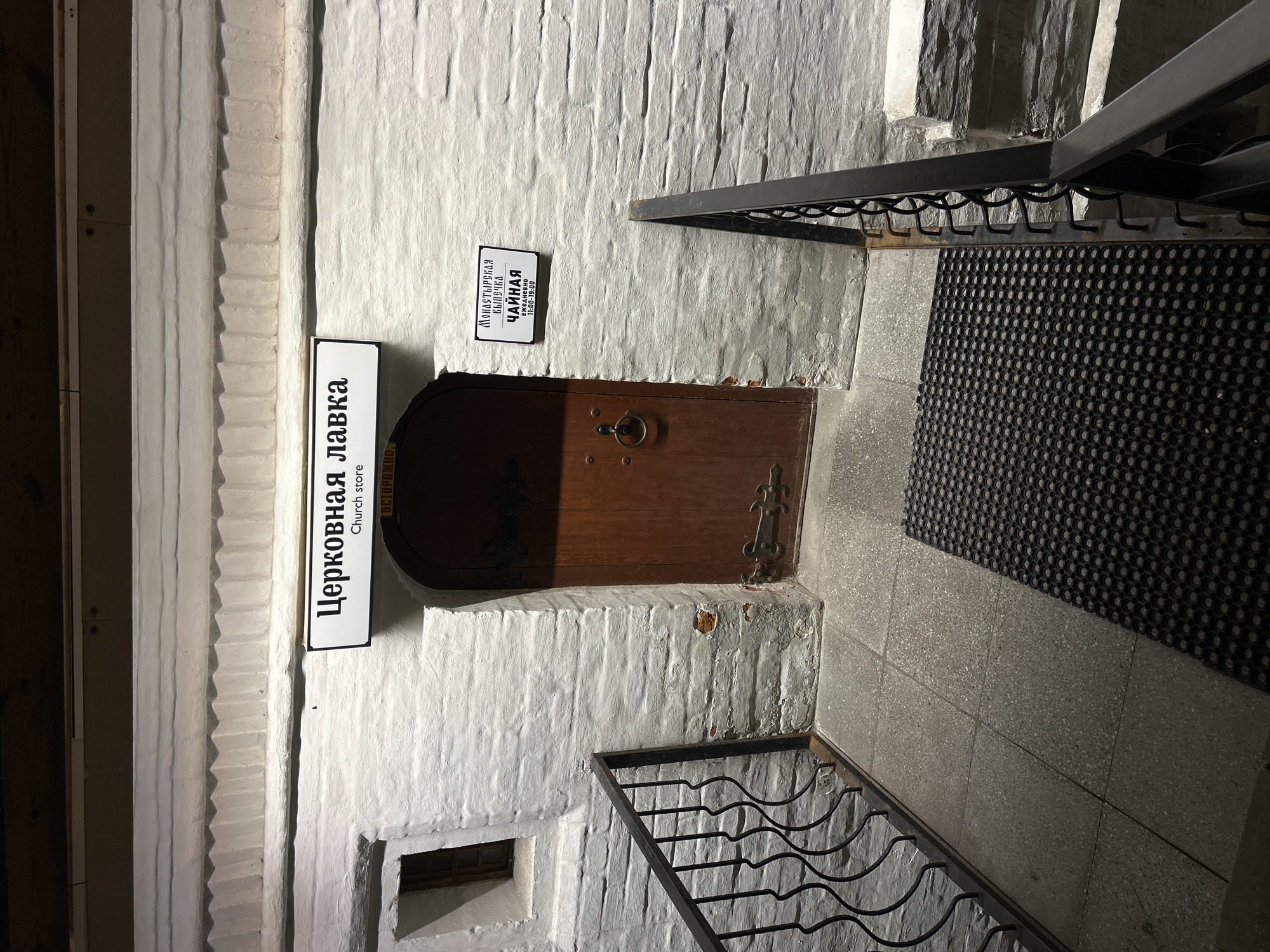 The image shows an exterior view of an entrance to a building in Moscow, Russia, at night. A white brick wall is visible, with a wooden door and two signs. The sign above the door reads "Церковная лавка" and "Church store". To the right of the door, another sign reads "МОНАСТЫРСКАЯ БЫЛЫЧКА" and "ЧАЙНАЯ", with operating hours listed below. A black metal railing leads up to the entrance. The ground is paved with square tiles and a black rubber mat. The lighting suggests it is nighttime.