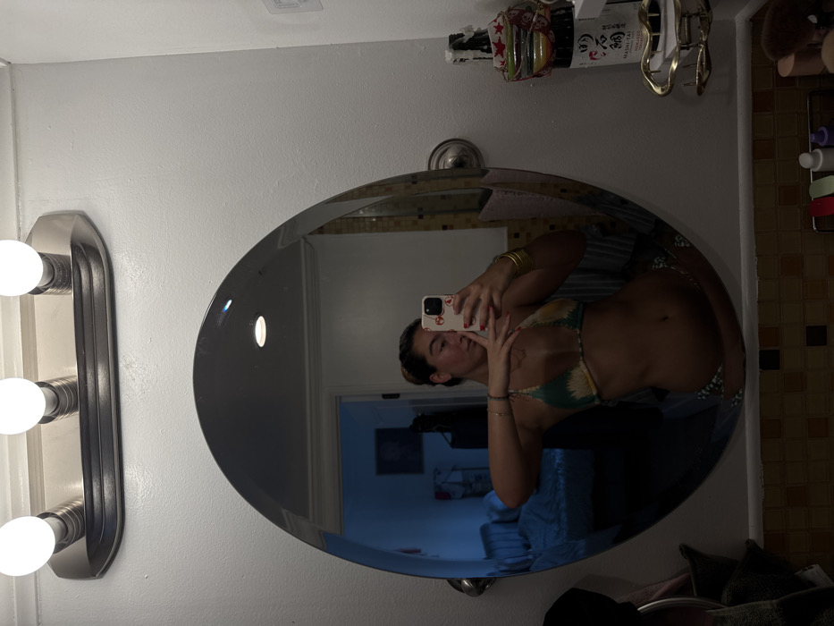The image displays an interior scene within a residential bathroom in the city of Los Angeles, United States of America. An adult female is reflected in a large, oval, wall-mounted mirror, engaged in the act of taking a selfie. She is wearing a green, yellow, and white patterned bikini top and matching bottoms. Her left hand holds a white smartphone with a red-patterned case, positioned to capture her reflection; her fingernails are painted red. Her right arm is extended laterally, adorned with multiple gold-colored bangles on the wrist, while a thin bracelet is visible on her left wrist. Distinct tan lines are visible across her upper chest, indicating prior sun exposure from a different garment.
The bathroom environment features white painted walls and a chrome light fixture with three exposed round bulbs mounted above the mirror. Below the mirror, a tiled surface composed of small square tiles in shades of brown, orange, and tan is visible. To the immediate right of the mirror, a multi-tiered clear plastic shelf unit holds various toiletry bottles. Additional items on the counter include a white bottle with Japanese script and a small clear container holding red and white star-shaped objects, situated above a gold-colored wire shelf bracket. A dark towel hangs to the left of the mirror.
Reflected in the mirror, behind the individual, is an adjacent room, likely a bedroom, discernible through an open doorway. This room contains a partially made bed with blue or grey bedding, other furniture, and a framed artwork hanging on a wall. The reflected room appears dimly lit with a blue hue. The exact address within Los Angeles is not identifiable from the visual information provided.