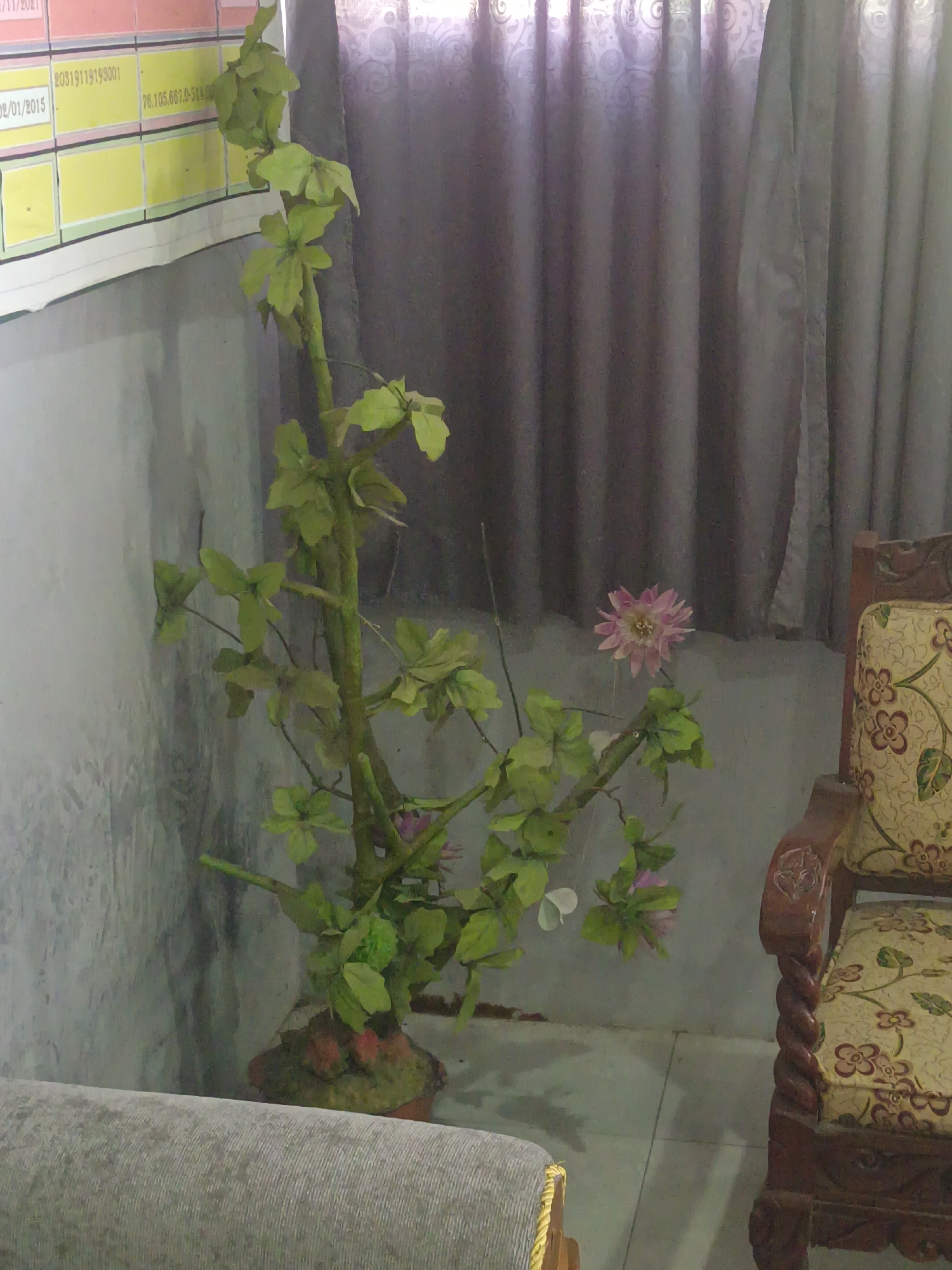 The image shows an artificial potted plant with green leaves and a pink flower in a room. To the left of the plant is a wall with a notice board or calendar. To the right of the plant is a wooden armchair with a floral pattern. The room appears to be indoors, with grey curtains in the background. The overall lighting suggests it could be daytime, but the curtains are drawn. There are no people visible in the image. The location is identified as Sarimulyo, Indonesia.