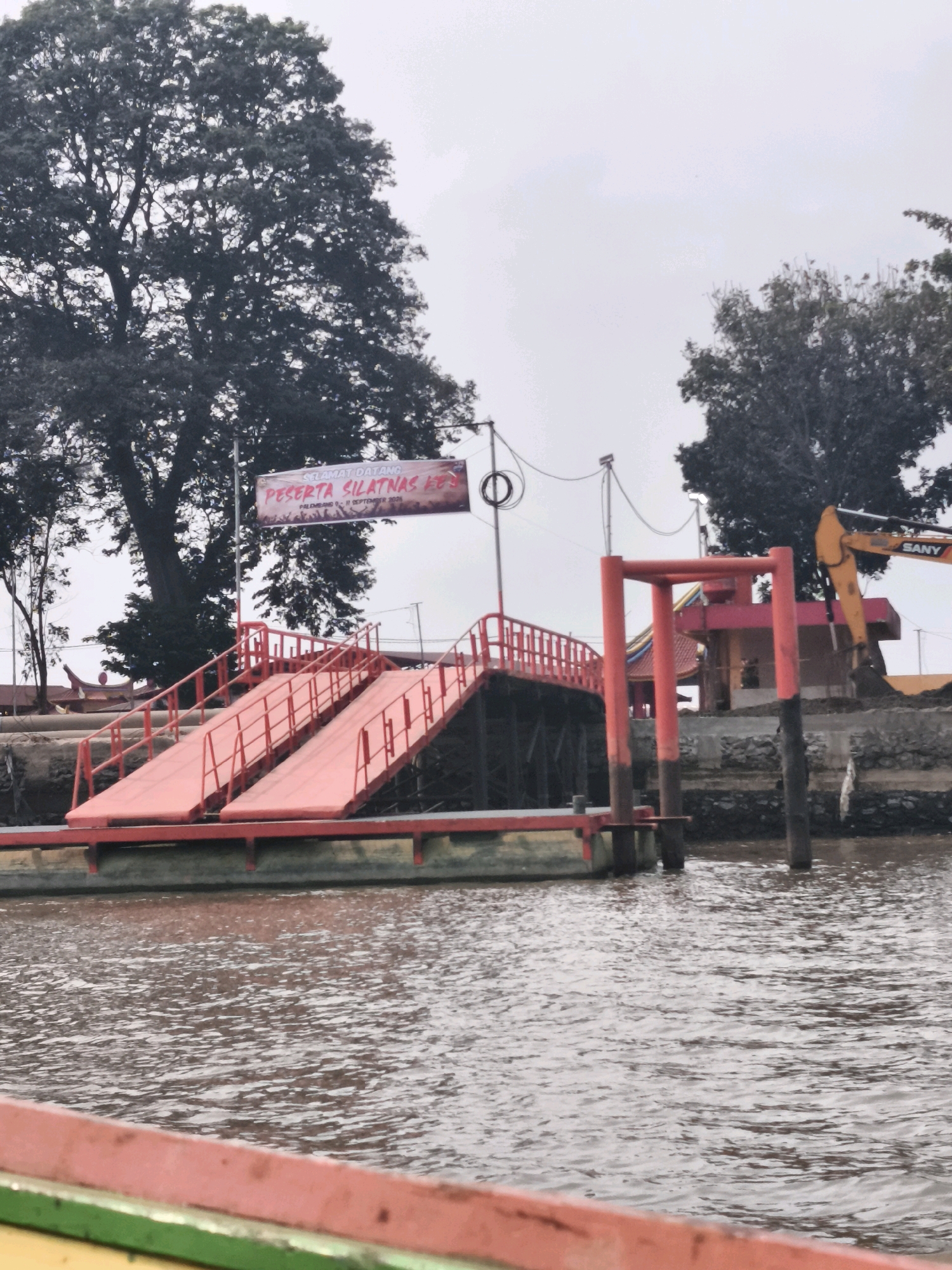 The image depicts a red ramp leading from a body of water to an elevated platform, possibly a ferry terminal. A banner above the ramp reads "SELAMAT DATANG PESERTA SILATNAS X" and "PALEMBANG 9-11 SEPTEMBER 2021". To the right, a yellow excavator is visible, suggesting ongoing construction or maintenance. In the background, lush trees and a hazy sky contribute to the ambiance. The scene is likely located in Palembang, Indonesia, given the text on the banner and the characteristic tropical vegetation.