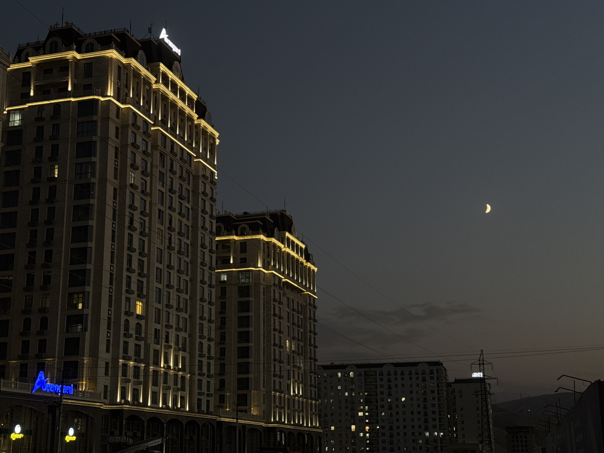 The image shows a twilight view of modern buildings in Bishkek, Kyrgyzstan. The prominent structure on the left is a tall, multi-story building with ornate architecture and illuminated gold trim along its upper levels and vertical accents. A prominent "Avangard" sign with a triangular logo is visible at the top of this building. To its right, a slightly shorter, similarly designed building is also illuminated. In the background, other apartment buildings with lit windows are visible, indicating residential activity. The sky is dark blue with a faint crescent moon and wispy clouds. Power lines are visible stretching across the upper portion of the scene. The overall impression is an urban landscape at dusk, with architectural illumination being a key feature.