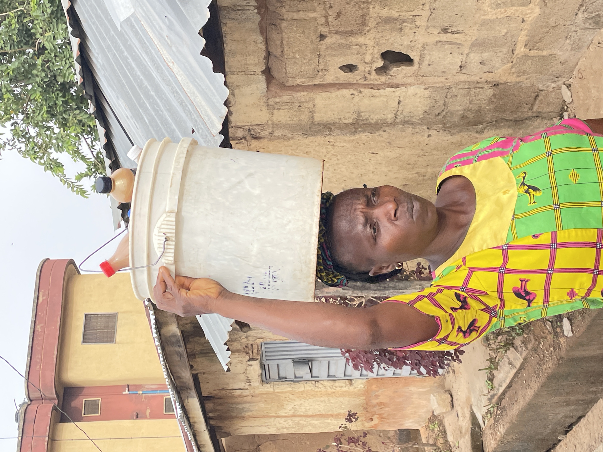 A woman stands outdoors in Jos, Nigeria, balancing a large, white plastic bucket on her head. The bucket is slightly tilted forward, with two plastic bottles, one brown and one red, suspended from its sides. She extends her right arm to support the bucket by holding its wire handle. She wears a colorful, patterned shirt with yellow, green, and pink squares and black bird designs. Her hair is tied back, and she has a serious expression. Behind her, there is a weathered building made of bricks and metal sheeting, possibly a home. Another building, painted in light yellow and red, is visible in the distance. The ground is earthy, with some vegetation growing nearby. The sky is overcast.