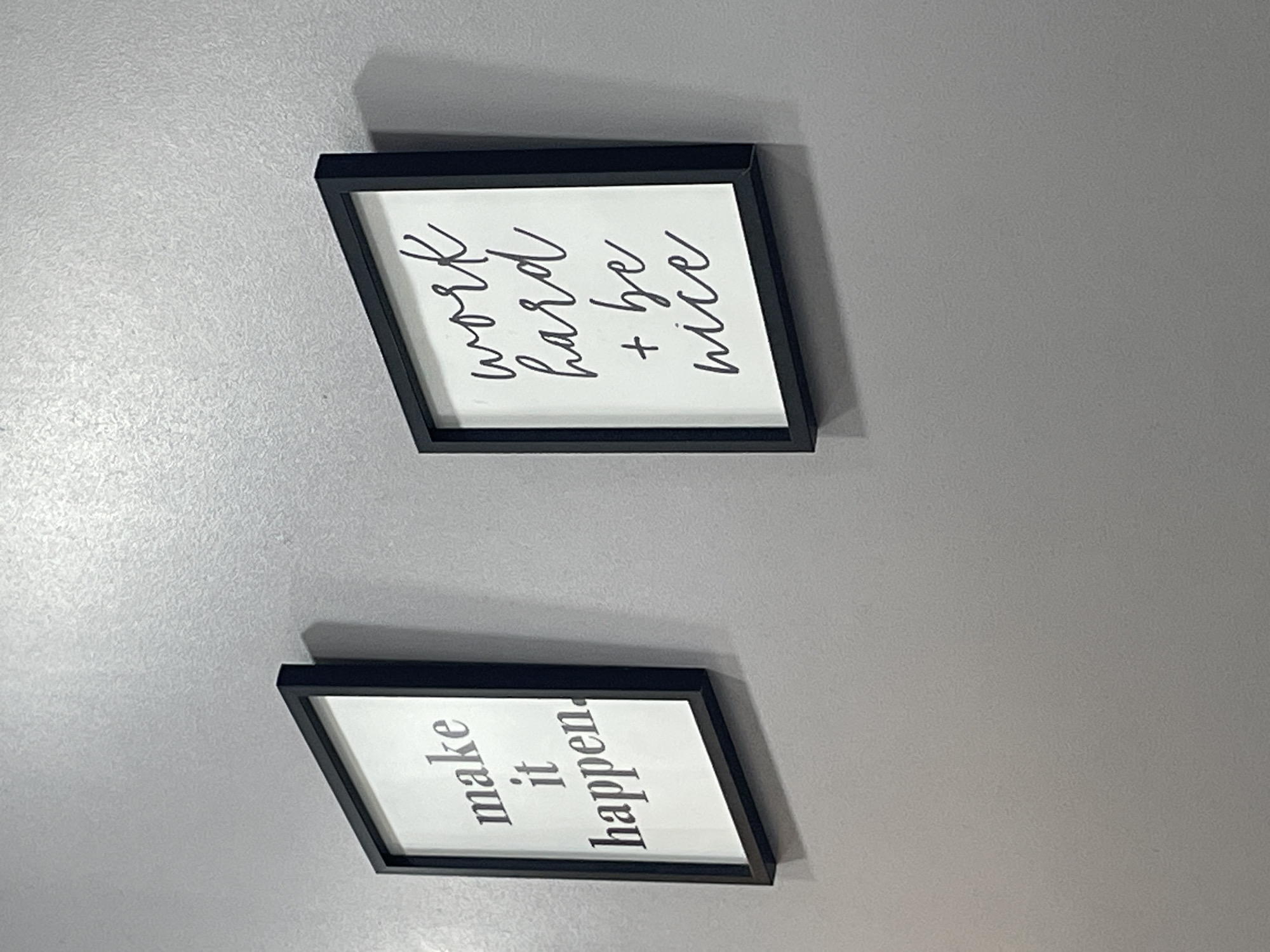 The image shows two framed prints hanging on a light gray wall. The top print reads "work hard + be nice" in black cursive lettering. The bottom print reads "make it happen" in black block lettering. The frames are black. The image is taken indoors. The city of Porlamar, Venezuela is not discernible from the image.