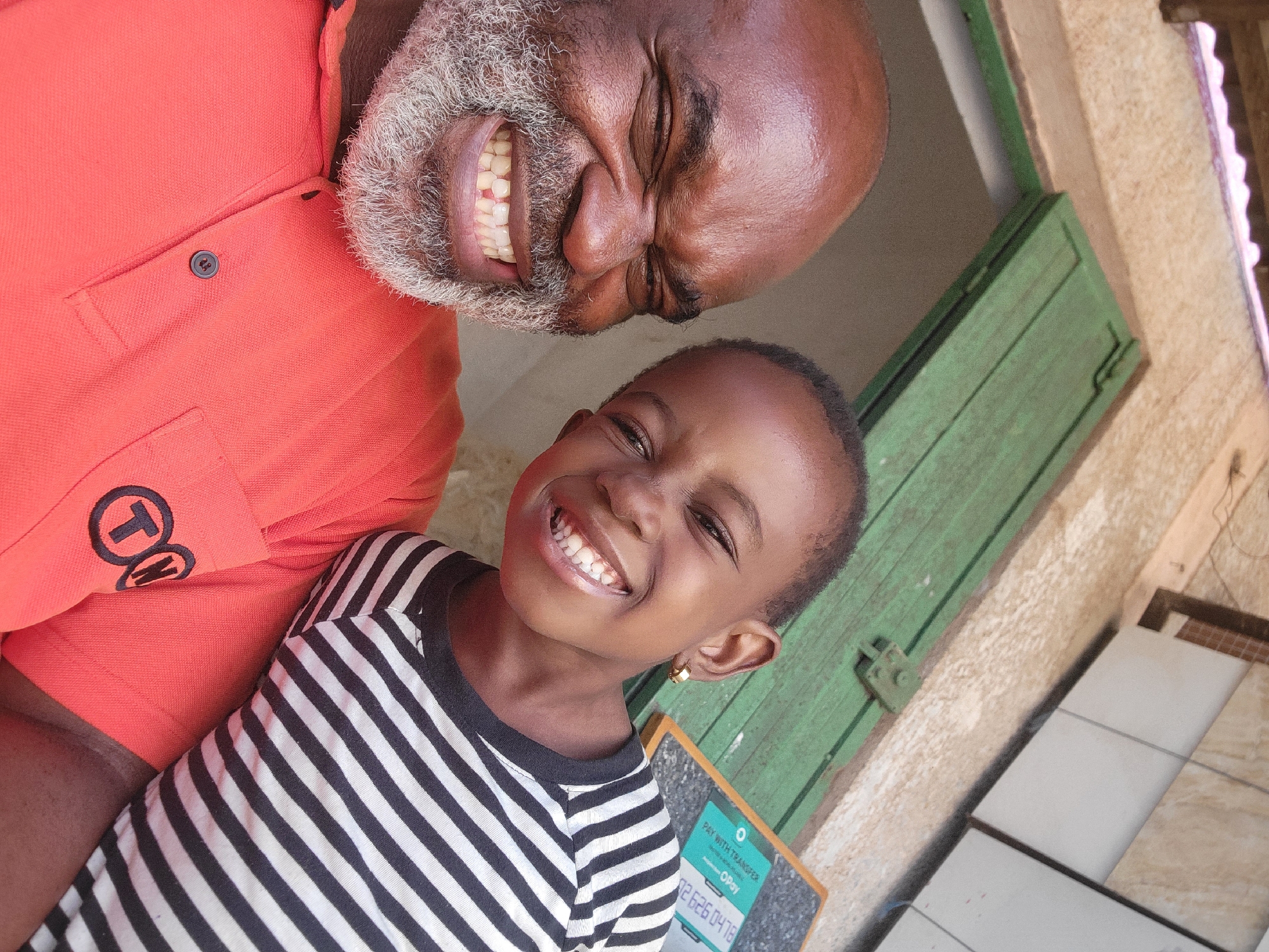 A man and a child are photographed together outdoors. The man is dark-skinned with a grey beard and is wearing an orange polo shirt with a logo on it. He is smiling widely, his eyes nearly closed. The child, also dark-skinned, is wearing a black and white striped shirt and is smiling as well. A green shuttered window is visible behind them. The child appears to be wearing a gold earring. A small signboard with numbers is partially visible near the child. Tiled construction materials are seen at the lower right of the photo.
The location is Maiburiji, Nigeria.