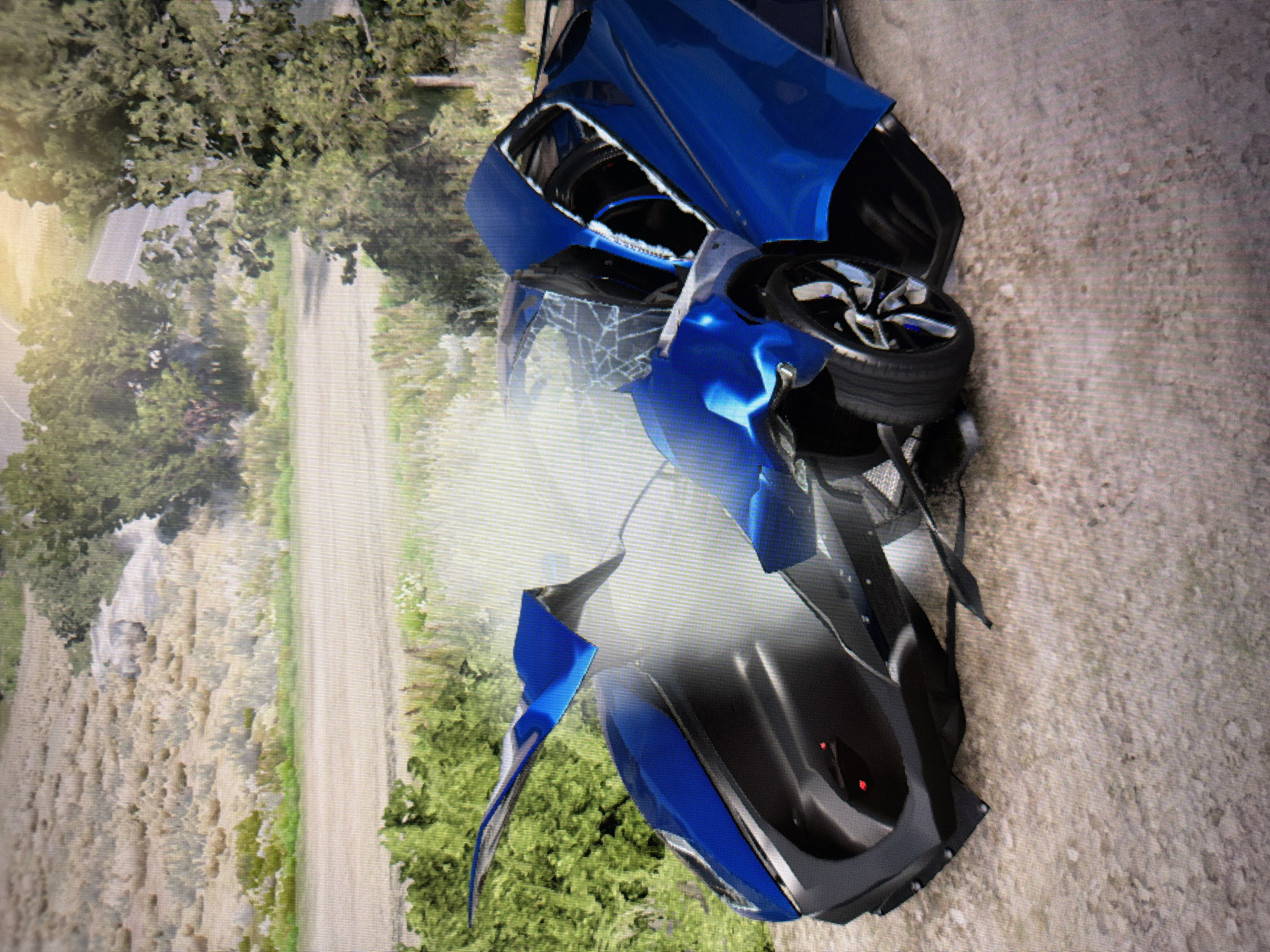 The image depicts a severely damaged blue sports car in what appears to be a rural or semi-rural outdoor setting. The car is situated on a dirt or gravel path, with lush green trees and bushes bordering the scene.
The front section of the car is extensively mangled and broken. The blue body panels are crumpled, torn, and displaced, revealing internal components and black accents that resemble carbon fiber. The windshield is severely shattered, exhibiting a distinct spiderweb crack pattern across its surface. A plume of white smoke or steam is seen rising from the front engine bay area, further indicating the impact of an accident. One of the car's wheels is visible, appearing askew and damaged, suggesting significant structural failure.
The background shows more trees and vegetation, along with what seems to be another unpaved road or track winding through the natural landscape. The lighting indicates it is daytime, with a bright, soft light source visible on the left side of the frame (if rotated upright), possibly suggesting morning or late afternoon. The overall texture and detail level of the image suggest it may be a screenshot from a video game rather than a photograph of a real-life event. No discernible text is visible anywhere in the image.