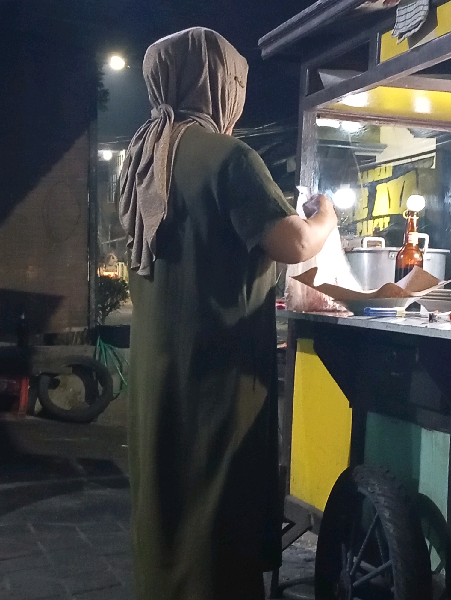 A woman wearing a headscarf and an olive green dress stands on a brick sidewalk next to a food stall at night. She is holding a white plastic bag open and appears to be placing items into it. The food stall is lit by overhead lights and features a yellow and green exterior. Various items are visible on the counter of the stall, including pots, a brown bottle, and paper wrapping. Two bicycle wheels are visible on the left and right sides of the image, and a green hose is coiled on the left. The image appears to have been taken in Kludan, Indonesia, based on the context provided.