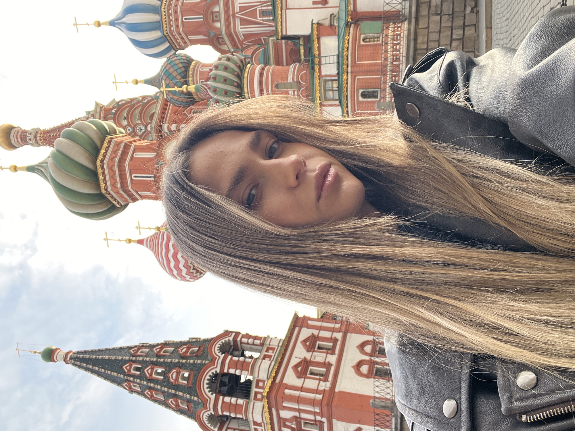 The image depicts a selfie of a female individual in front of St. Basil's Cathedral in Red Square, Moscow, Russia. The young adult female, positioned in the foreground, has long, straight blonde hair and is wearing a black leather jacket with silver-toned hardware. She is looking directly into the camera with a neutral expression. Behind her, St. Basil's Cathedral is prominently featured, characterized by its red brick architecture with white detailing and a multitude of colorful, distinctively patterned onion domes, including striped green, red, blue, and multi-colored designs, each topped with a golden Orthodox cross. A tall, dark-tiled tented roof structure with a green dome and cross is visible on the left side of the cathedral. The scene is outdoors under a partly cloudy sky with bright daylight. Elements of a paved ground and metal railing are partially visible in the lower sections of the image.
