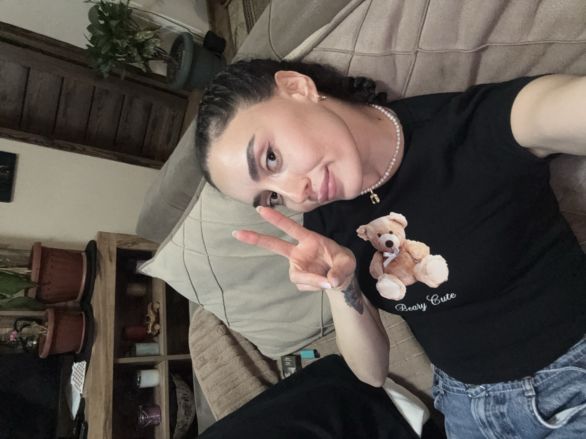 A woman is lying on a beige couch indoors, giving a peace sign. She is wearing a black t-shirt with a teddy bear design, a pearl necklace, and blue jeans. The setting includes wooden shelves with potted plants and other miscellaneous items. The location is Tbilisi, Georgia.