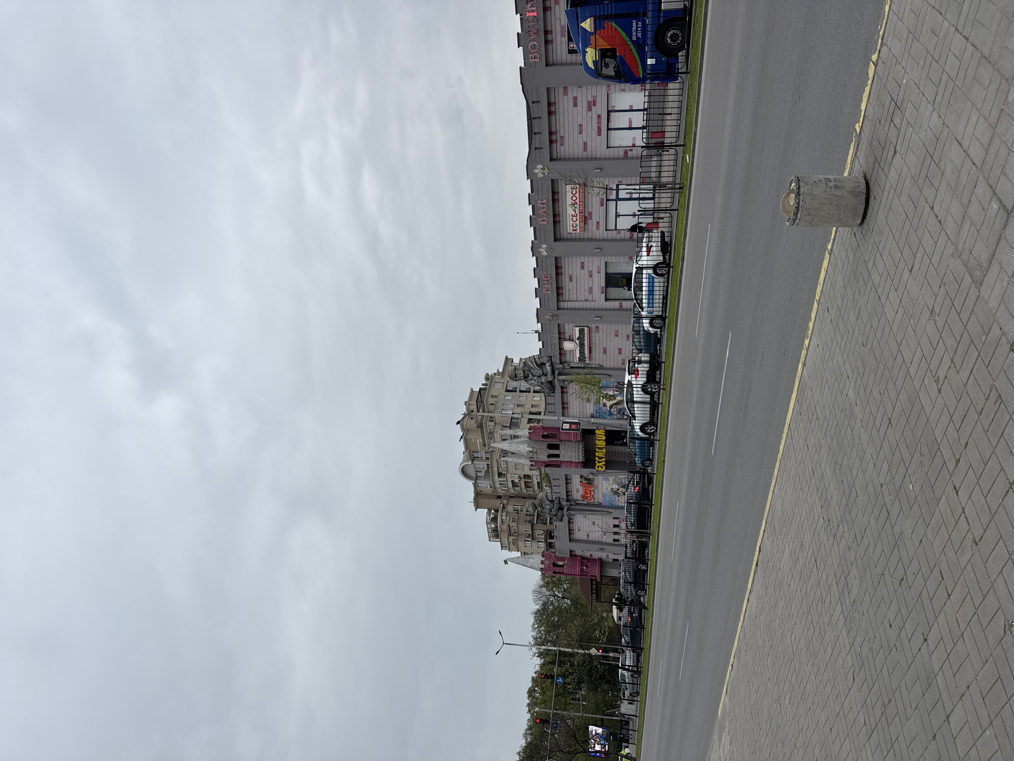 The image captures a street scene in Plovdiv, Bulgaria. On the right is a wide, grey brick sidewalk with a cylindrical concrete bollard at the edge. Adjacent to the sidewalk is a grey asphalt road with double yellow center lines. A large building with castle-like architecture dominates the left side of the image. It appears to be a commercial complex, with different sections possibly housing shops and restaurants. The building features multiple levels, turrets, and decorative elements. Banners and signage are visible on the building's facade. Parked cars line the street in front of the building, separated from the sidewalk by a black metal fence. Trees and foliage are present at street level in front of the castle-like building. The sky is overcast, suggesting a grey or cloudy day.
