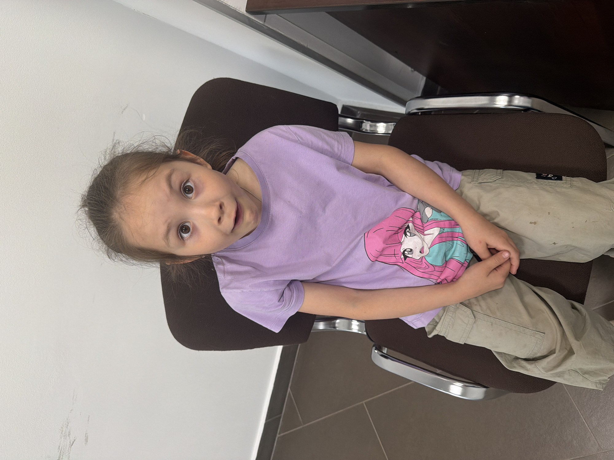A young girl is sitting in a brown chair with silver armrests. She is wearing a purple t-shirt with a cartoon character on it and khaki pants. She is looking up with a surprised expression. The background includes a white wall, a dark brown surface, and a tiled floor. The location is identified as Perm, Russia.