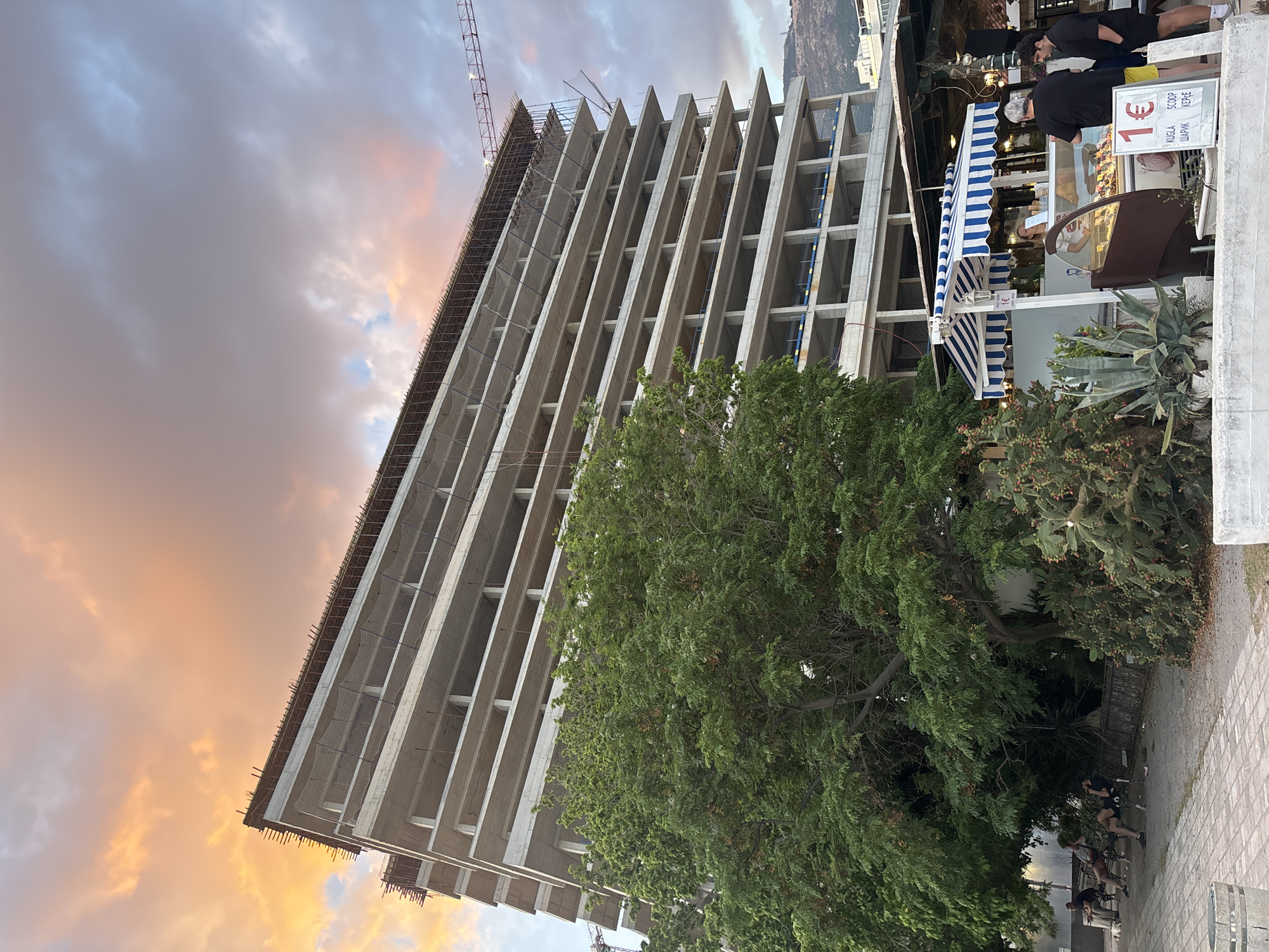 The image depicts an unfinished concrete building in Budva, Montenegro, at sunset. The building's framework is visible, with rebar extending from the top floor. A construction crane is present on the roof. A large tree partially obscures the lower floors. To the right, a striped blue and white awning covers an ice cream stand, where two people are being served. In the lower right corner, a group of people is visible under the tree. The sky is filled with clouds colored orange and gray by the setting sun. A sign indicates ice cream is €1 per scoop.