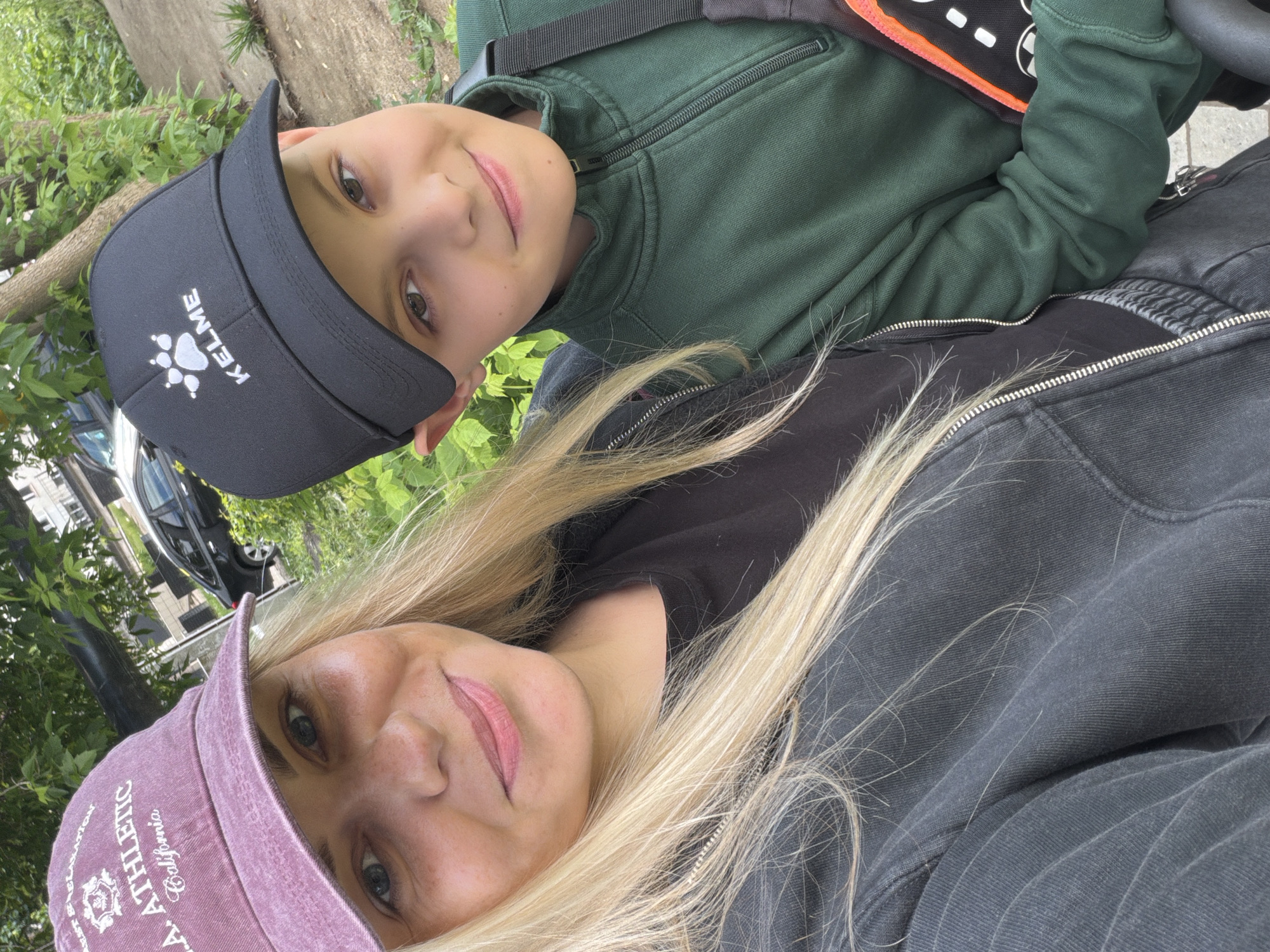 An outdoor, rotated selfie captures an adult and a child smiling directly at the camera. The image is oriented sideways, with the subjects' heads positioned to the left.
The adult, likely a woman, is in the foreground. She has long, blonde hair falling over her shoulders and is wearing a dark top or jacket. On her head is a dusty pink baseball cap with white embroidered text. Visible words include "ATHLETIC", "REGULATION", "California", and partially "U.S.A.". Her expression is a gentle smile.
Behind her and slightly above, the child, likely a boy, is also smiling. He wears a dark green zip-up hoodie, a black baseball cap with a white logo, and a visible strap of an orange and black backpack over his shoulder. The logo on his cap is a paw print, above the word "KELME".
The background features lush green foliage, suggesting a park or tree-lined area. Parts of a dark-colored car and a building are visible behind the greenery on the left side of the frame. The overall mood conveyed is happy and casual, typical of a daytime outdoor snapshot in Votkinsk, Russia.