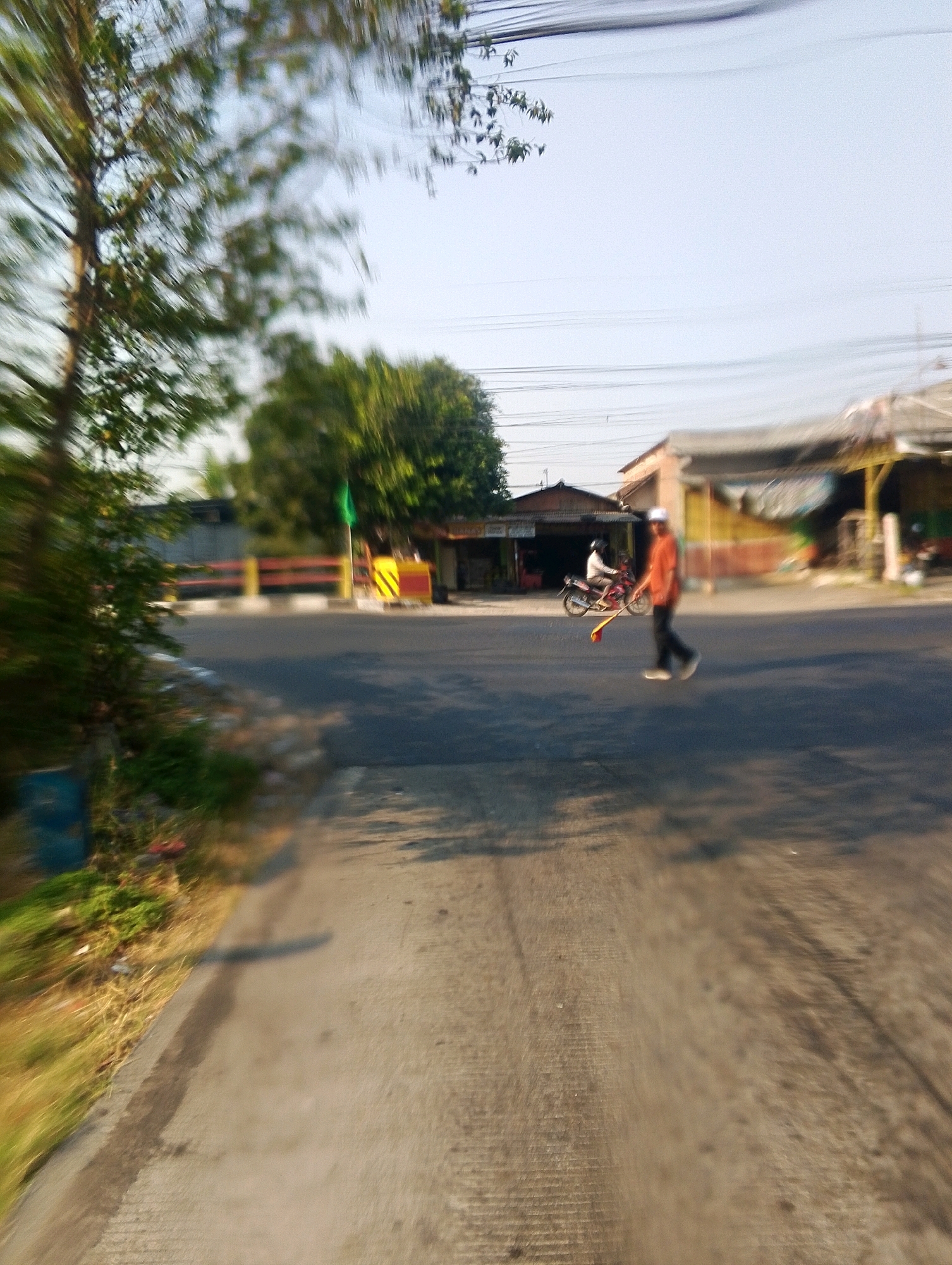 In an outdoor scene in Unknown, Unknown, a person wearing a white cap and an orange shirt is walking across a paved road. They are holding a yellow object with a red flag attached to it. The person is in motion, with their legs blurred. Behind them, a motorcycle is parked. In the background, there are several buildings with awnings and a green flag planted on a post. Trees are visible on the left and right sides of the image, with power lines overhead. The road appears to be an intersection with a yellow and red striped barrier. The overall image is blurry, suggesting movement or a low-quality camera.