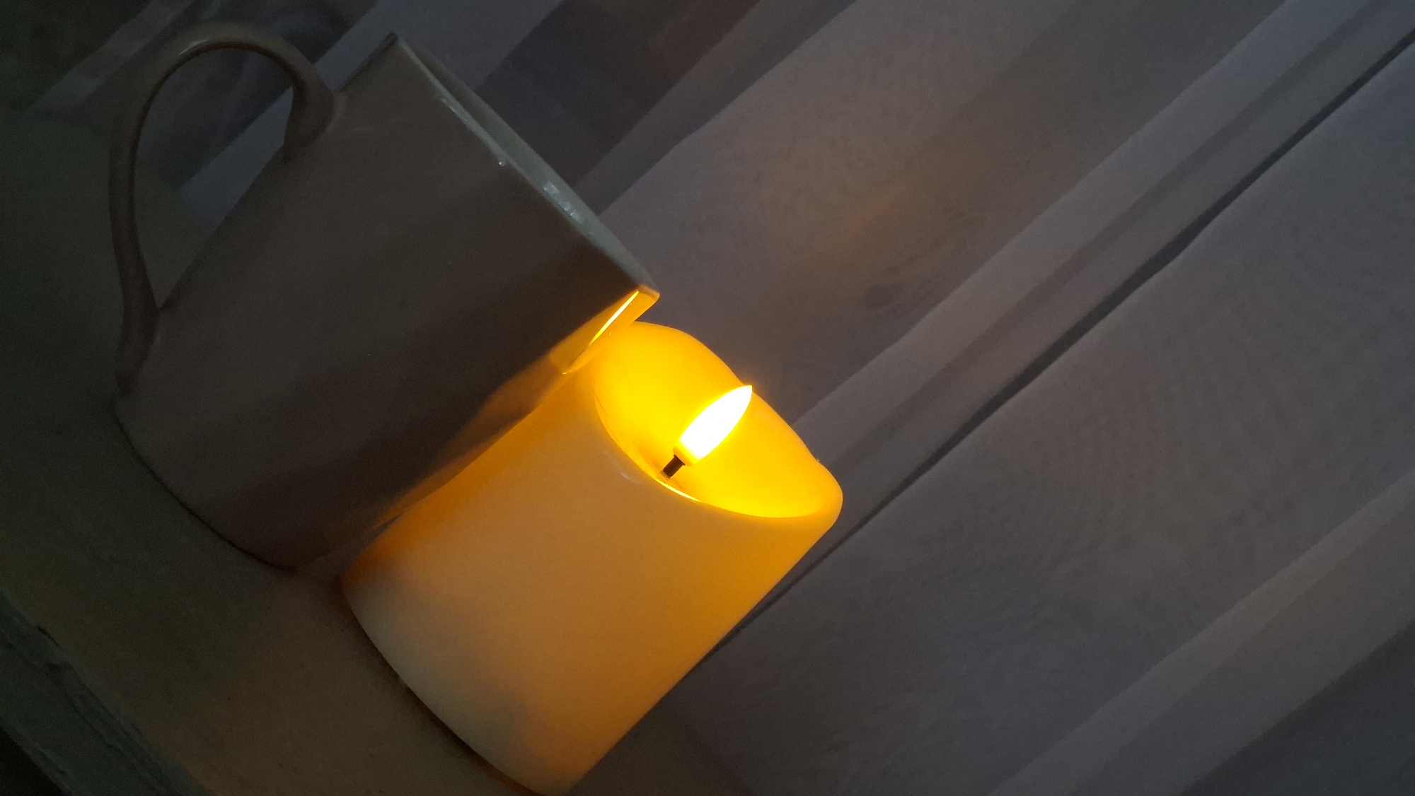 A lit candle is placed next to a mug on a surface, with pleated fabric in the background. The candle emits a warm yellow glow, illuminating the immediate surroundings. The mug is a light color and appears to be ceramic. The fabric in the background is light-colored and arranged in vertical pleats, suggesting it might be a curtain or drapery. The overall scene is dimly lit, with the candle being the primary light source. The image does not contain any information that would allow for its location in Myadzyel, Belarus, or any other specific geographic location.